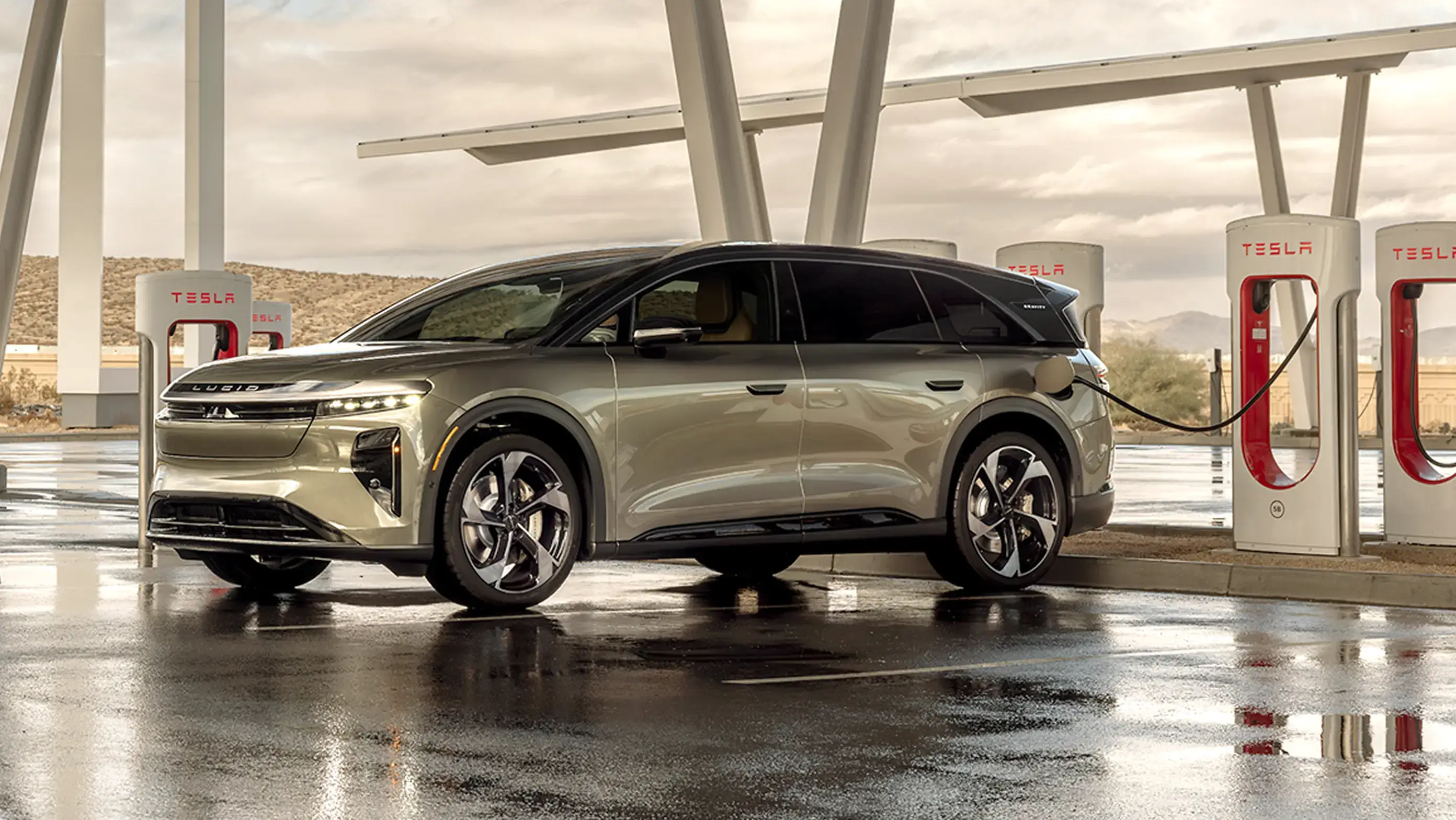 Lucid Gravity SUV charging at a Tesla Supercharger station under a modern canopy on a wet, cloudy day.