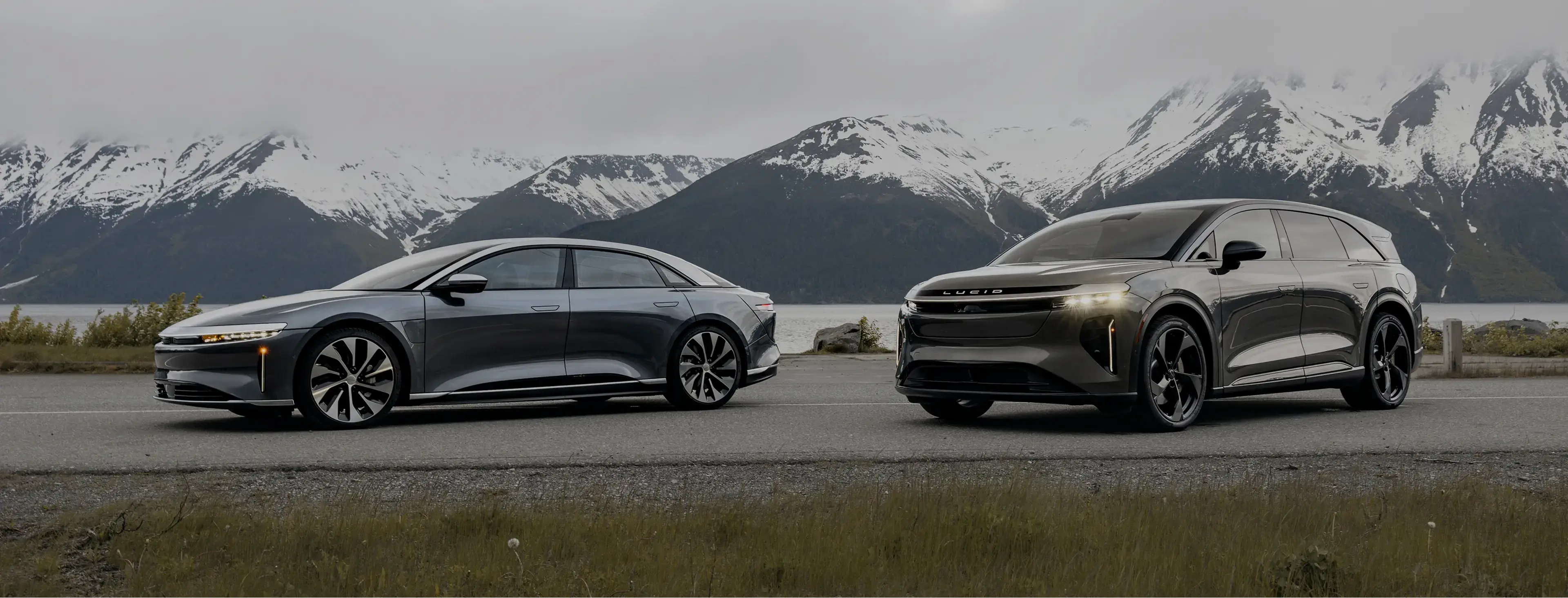 Two sleek Lucid electric vehicles parked on a road with snow-capped mountains and water in the background.