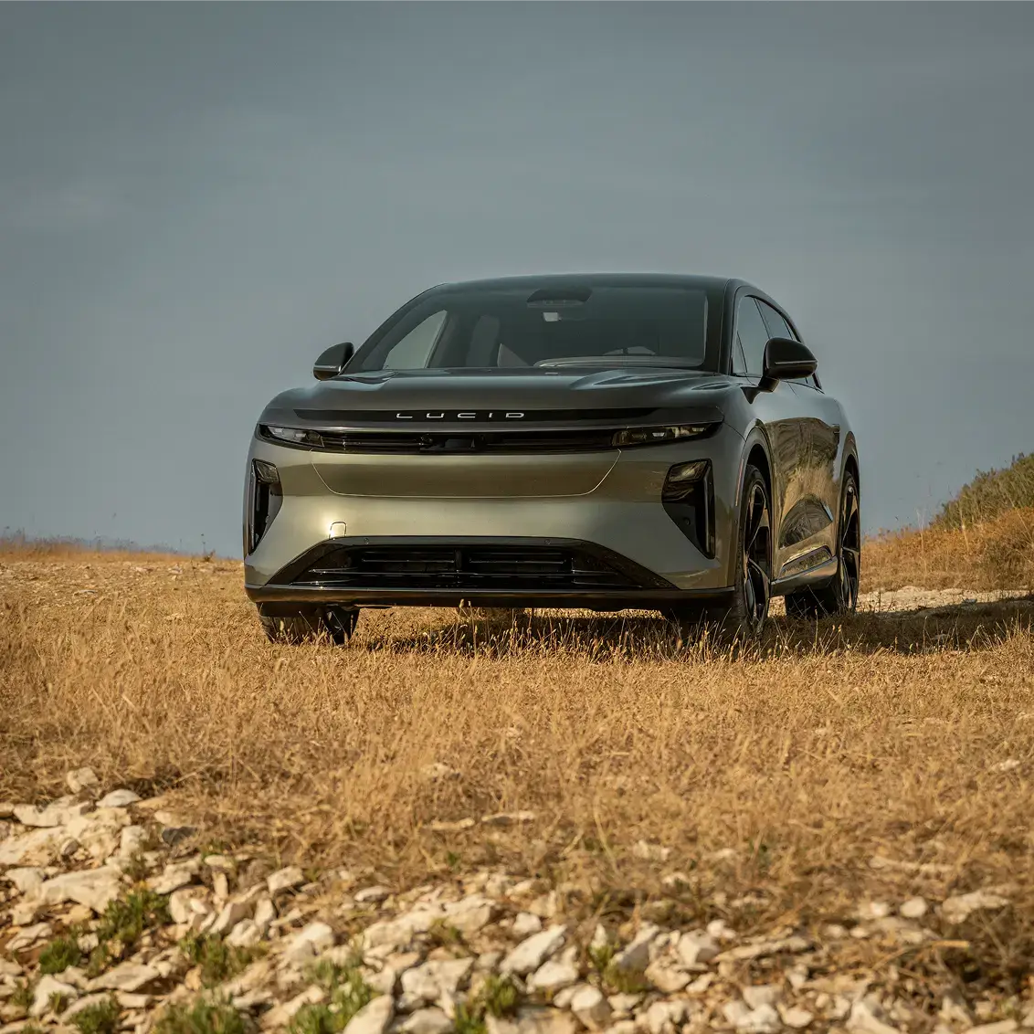 A silver Lucid electric SUV parked on a dry, rocky field under a gray sky, viewed from the front.