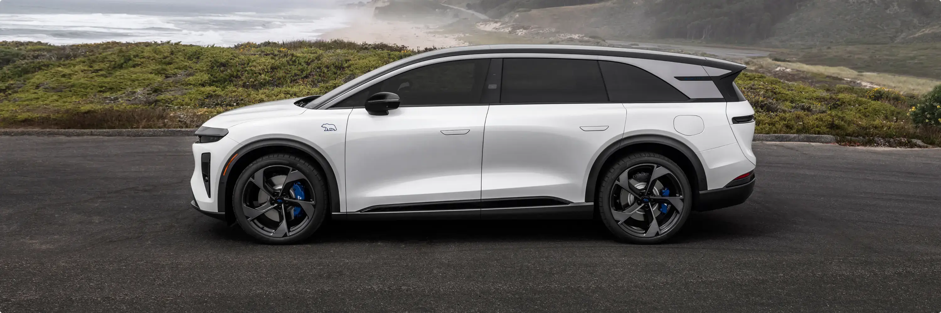 White electric SUV with black roof and blue brake calipers parked on a coastal road with misty ocean and green hills.