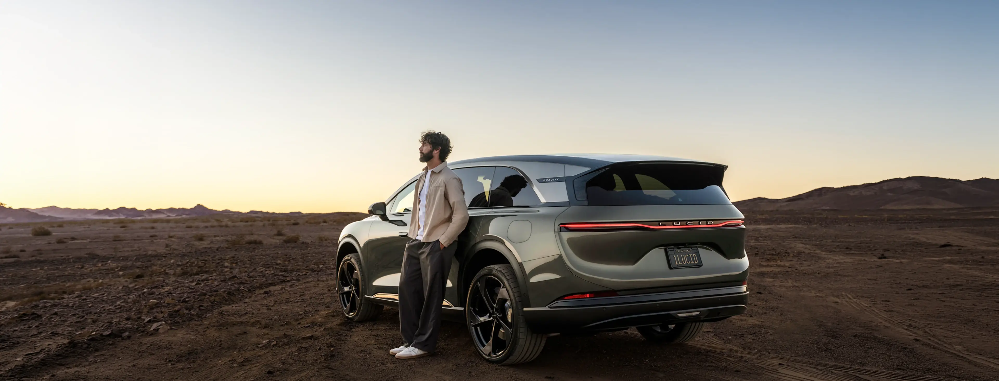 A man leans against a gray Lucid Gravity SUV in a desert landscape at sunset, gazing into the distance.