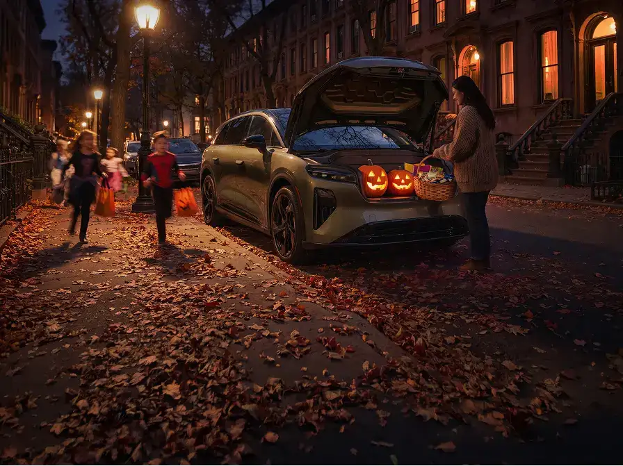 A woman holds a candy basket at an open car trunk with glowing jack-o'-lanterns as costumed children approach on a leaf-covered street.
