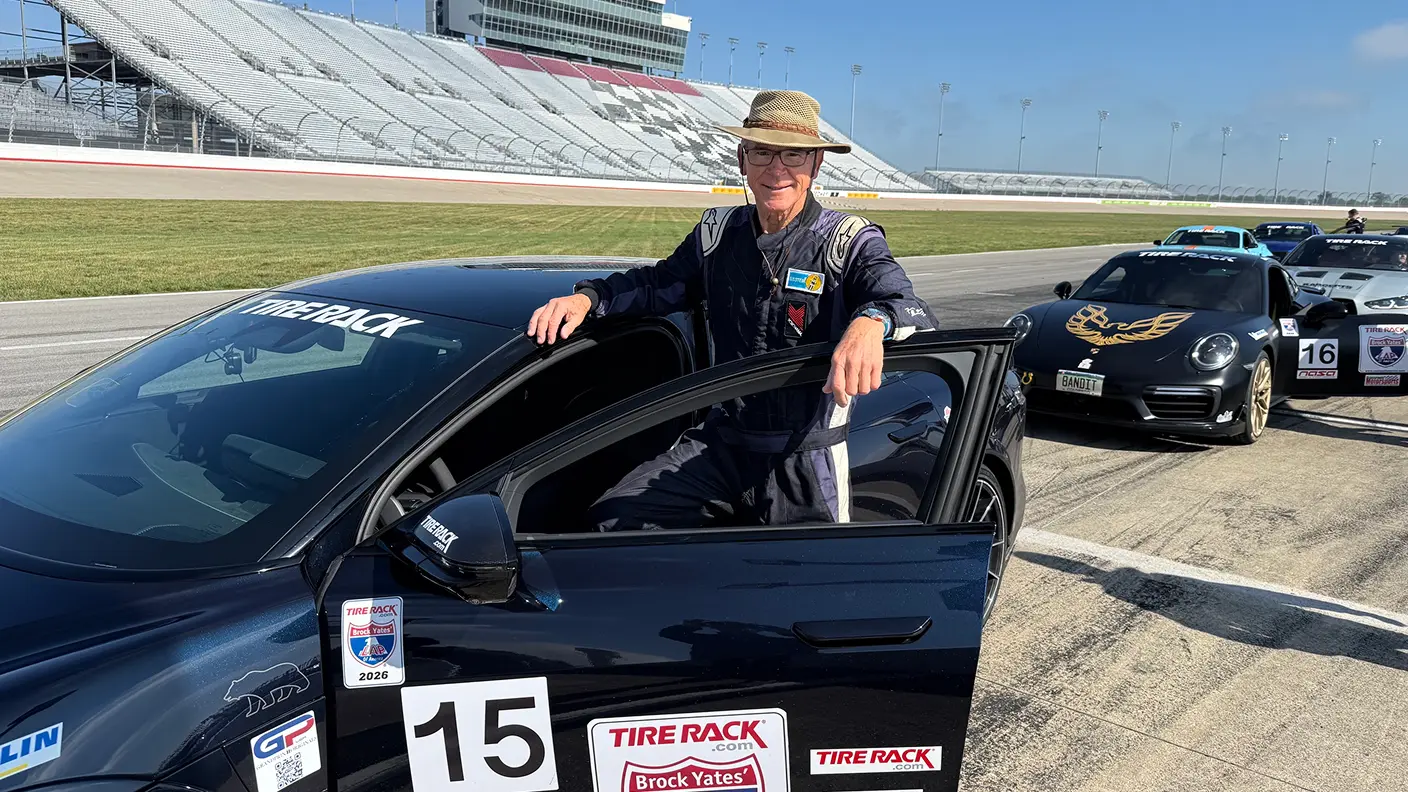 An older man in a racing suit stands beside a Lucid Air Sapphire numbered 15 at a racetrack, with grandstands and other race cars behind him.