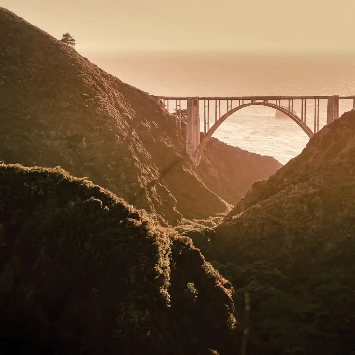 Bixby Bridge spanning a coastal canyon at sunset, with golden light illuminating rugged cliffs and ocean horizon.
