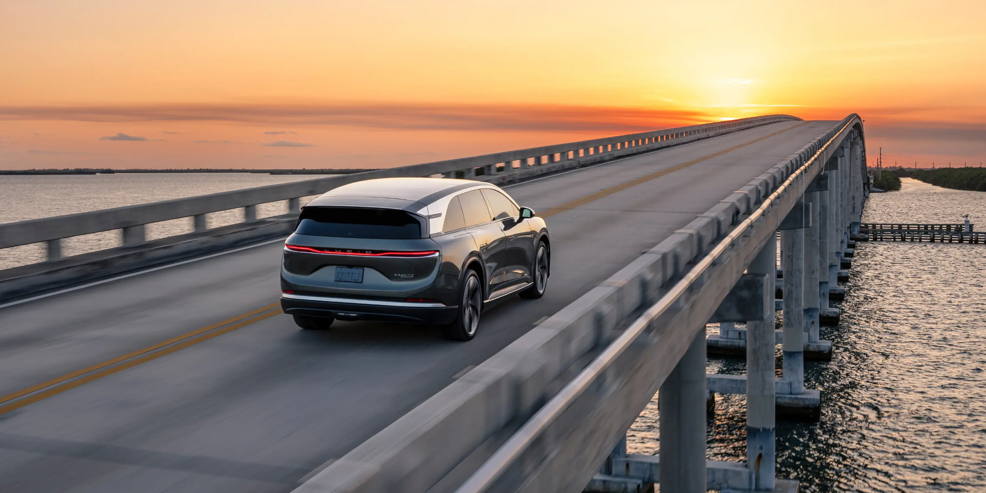 A Lucid Gravity SUV drives over a long bridge above water beneath a vibrant orange and yellow sunset sky.