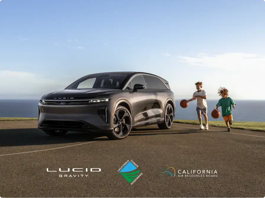 Lucid Gravity electric SUV parked outdoors near ocean, two children playing basketball nearby, sunny sky.