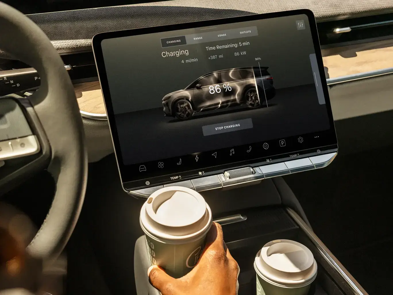 Electric vehicle dashboard showing 86% charge with 5 minutes remaining, while a hand holds a coffee cup in the cupholder.