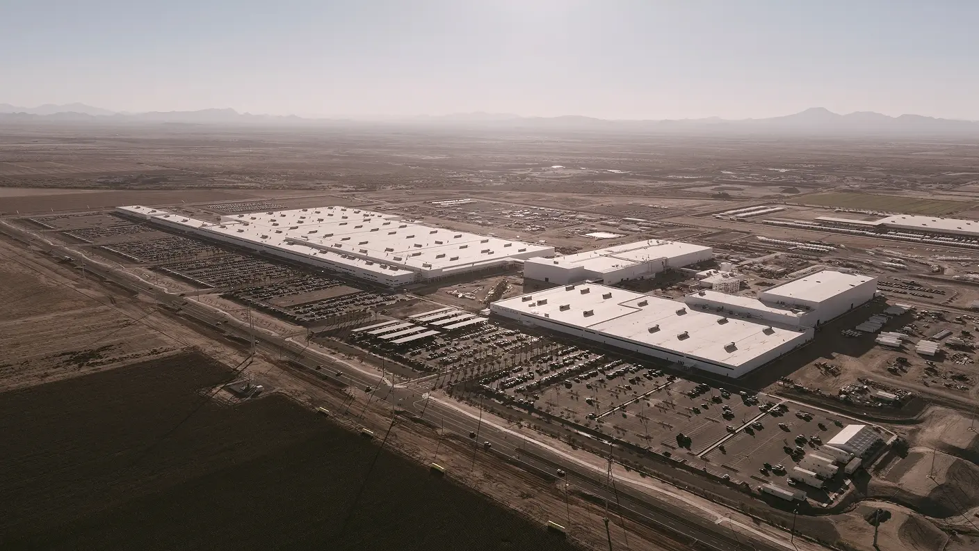 Aerial view of large industrial complex with white warehouse buildings and parking lots in a desert landscape with mountains in distance.