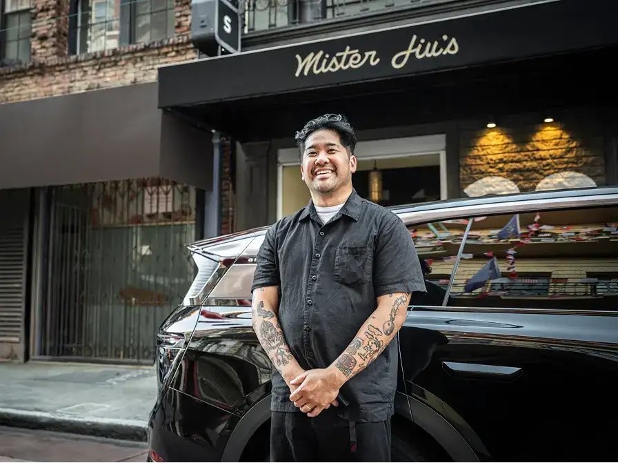 A smiling tattooed man in a dark shirt stands beside a black car in front of Mister Jiu's restaurant.