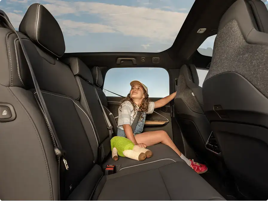 A young girl in overalls and a cap sits in a dark SUV back seat, looking up through a panoramic sunroof at the sky.