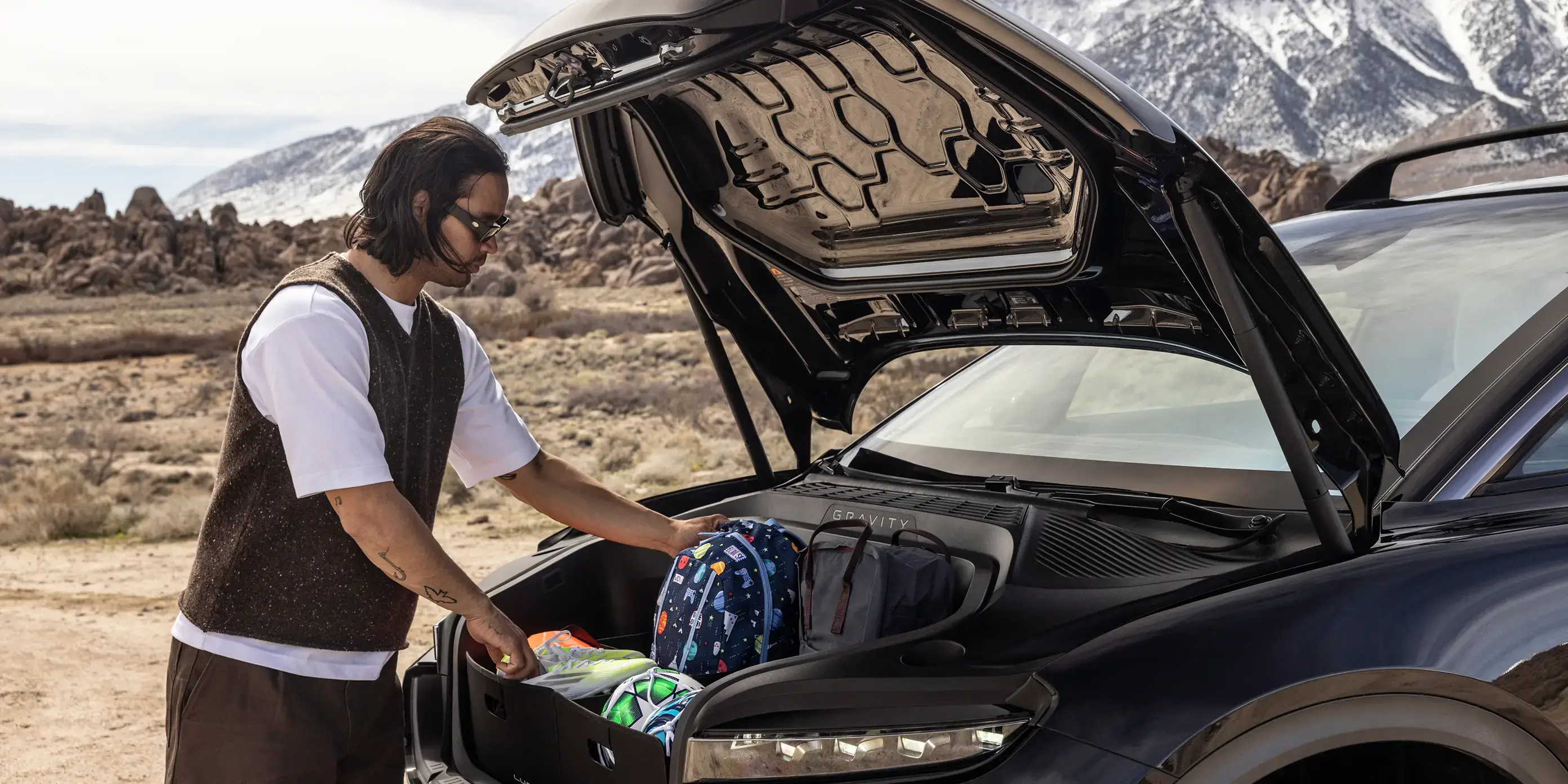 Person loading luggage into front trunk of black electric vehicle with snow-capped mountains in background.