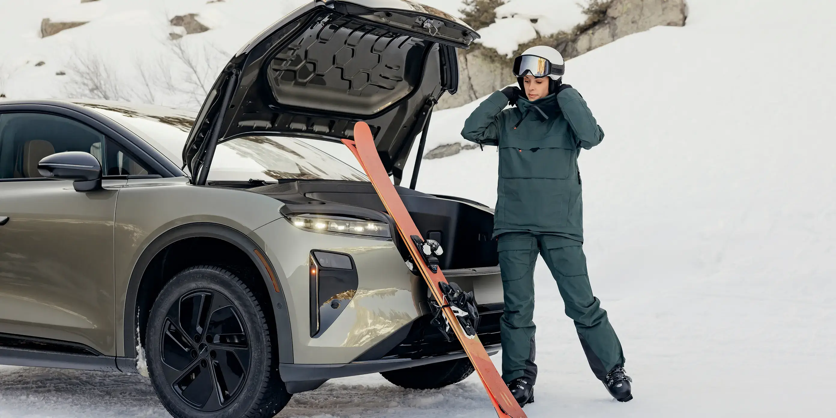 Un skieur en tenue de couleur verte ajuste son casque à côté d’un VUS Lucid Gravity avec coffre ouvert et des skis dans un décor de montagne enneigée.