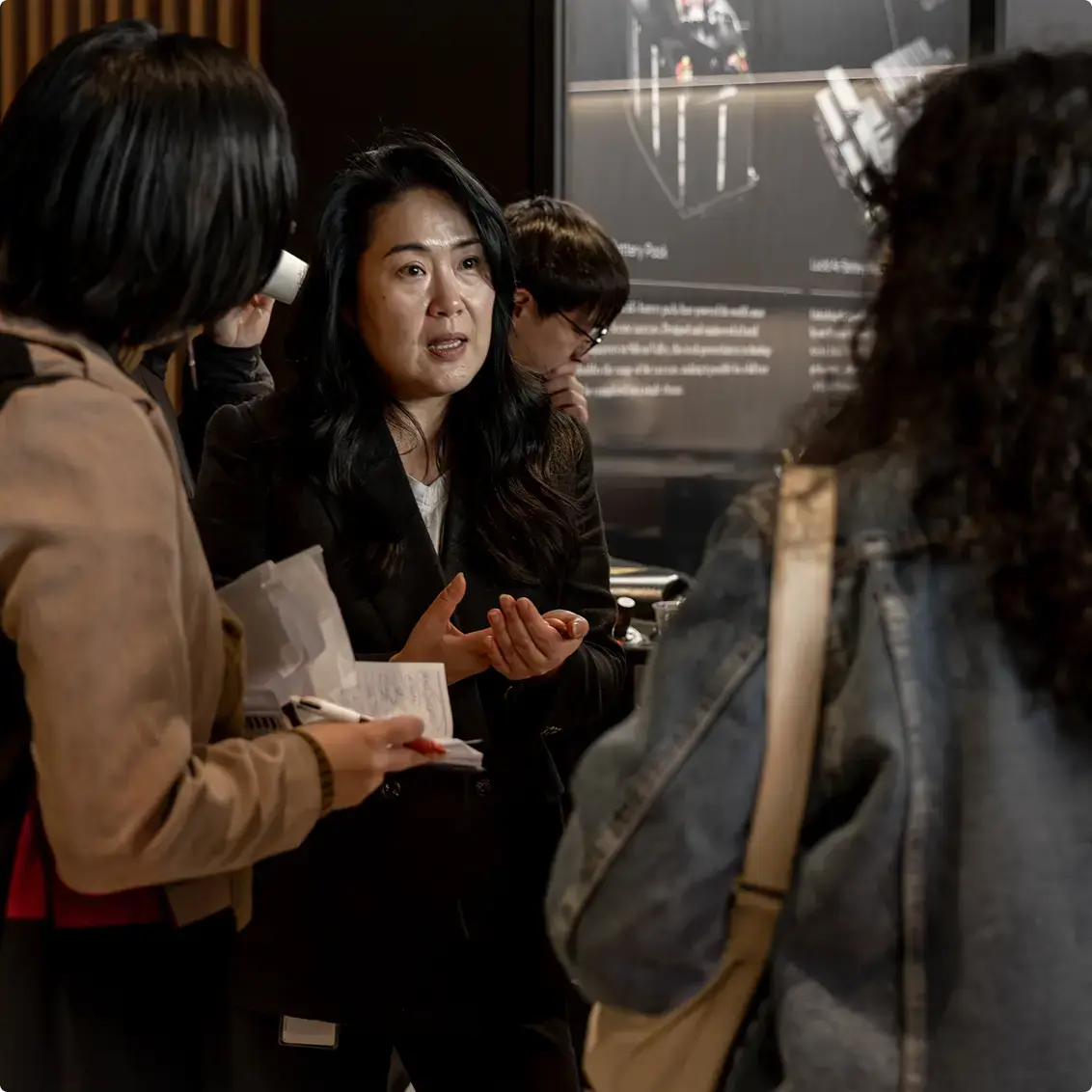 A woman in a black jacket gestures while speaking to colleagues during a presentation, with a digital display visible in the background.
