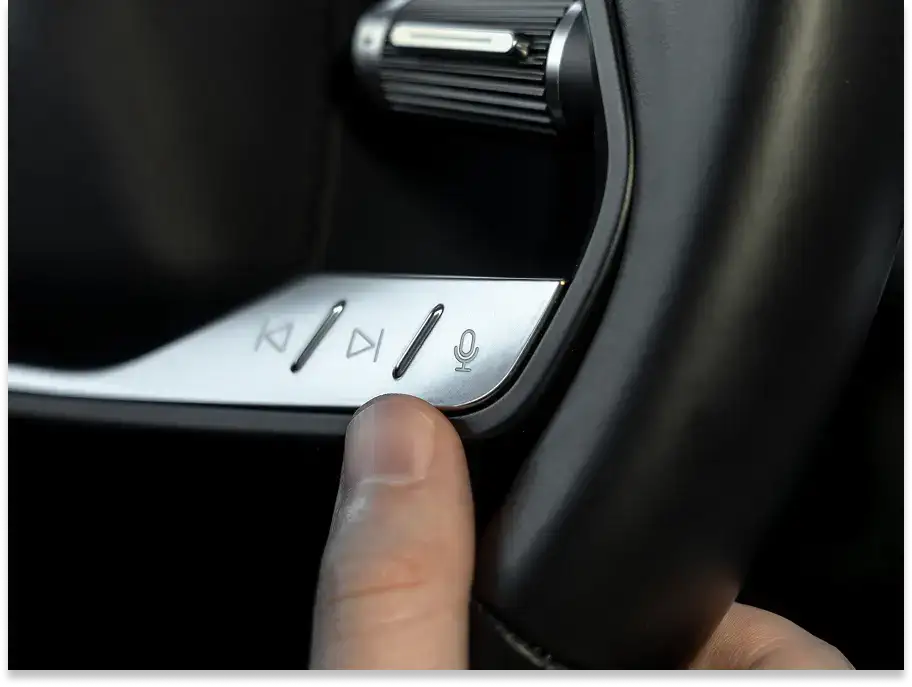 Finger pressing voice control button on car steering wheel with media controls and rotary dial visible.