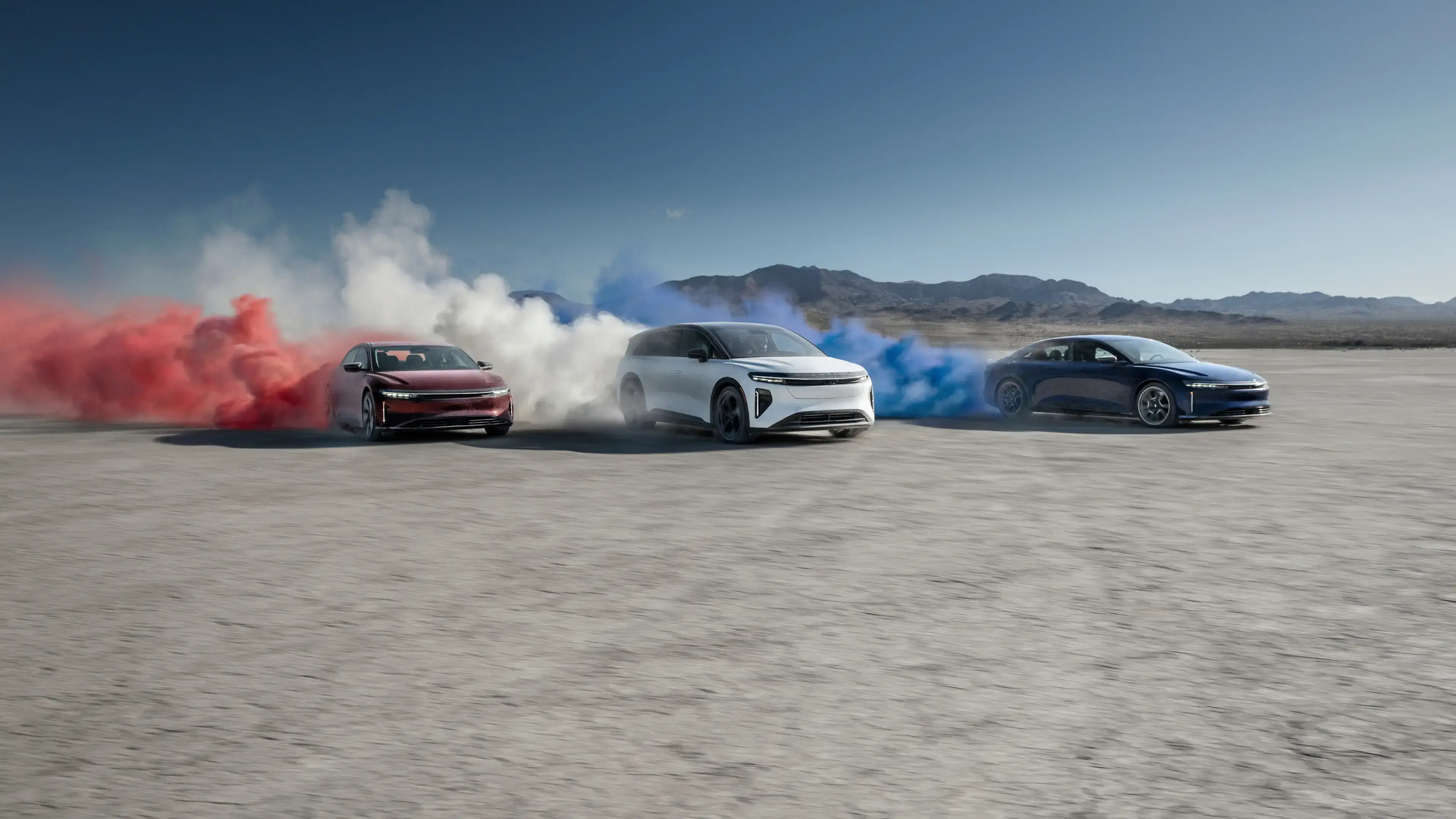 Three Lucid vehicles on a desert flat, trailing red, white, and blue smoke clouds under a clear blue sky.