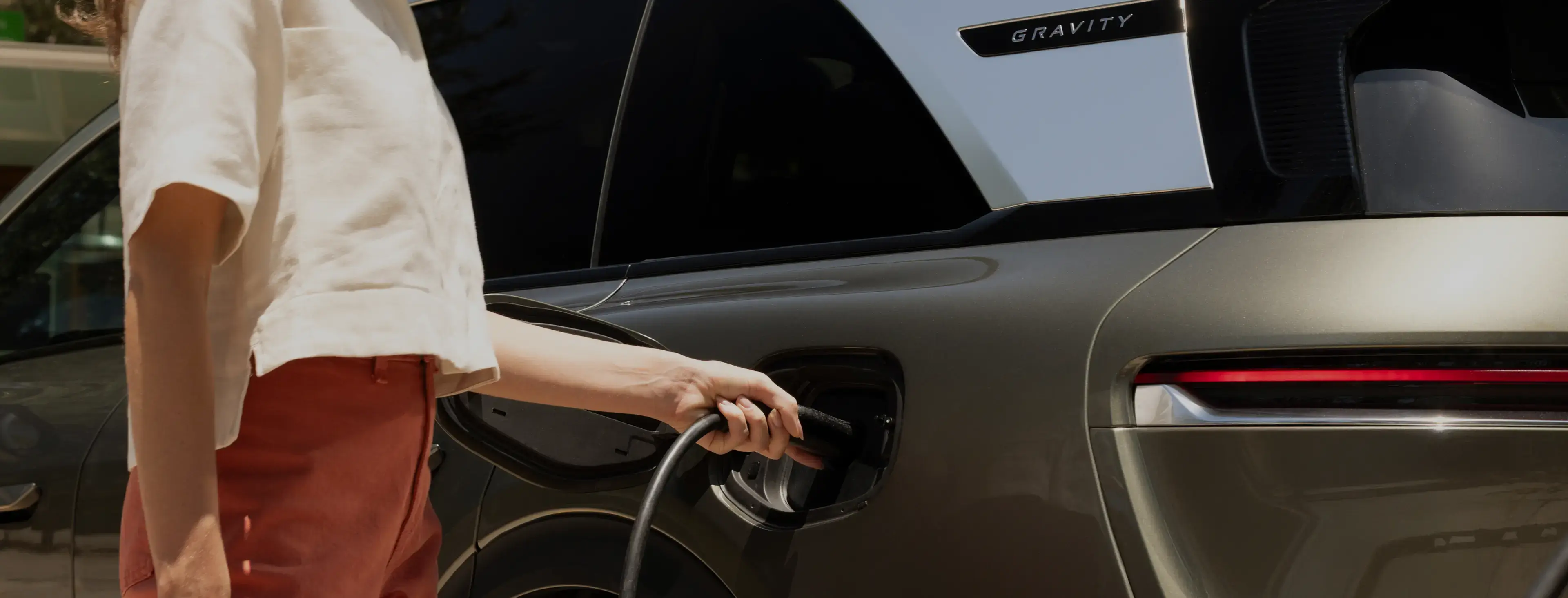 Person in white top and orange pants plugging in a charging cable to a Lucid Gravity electric vehicle outdoors.
