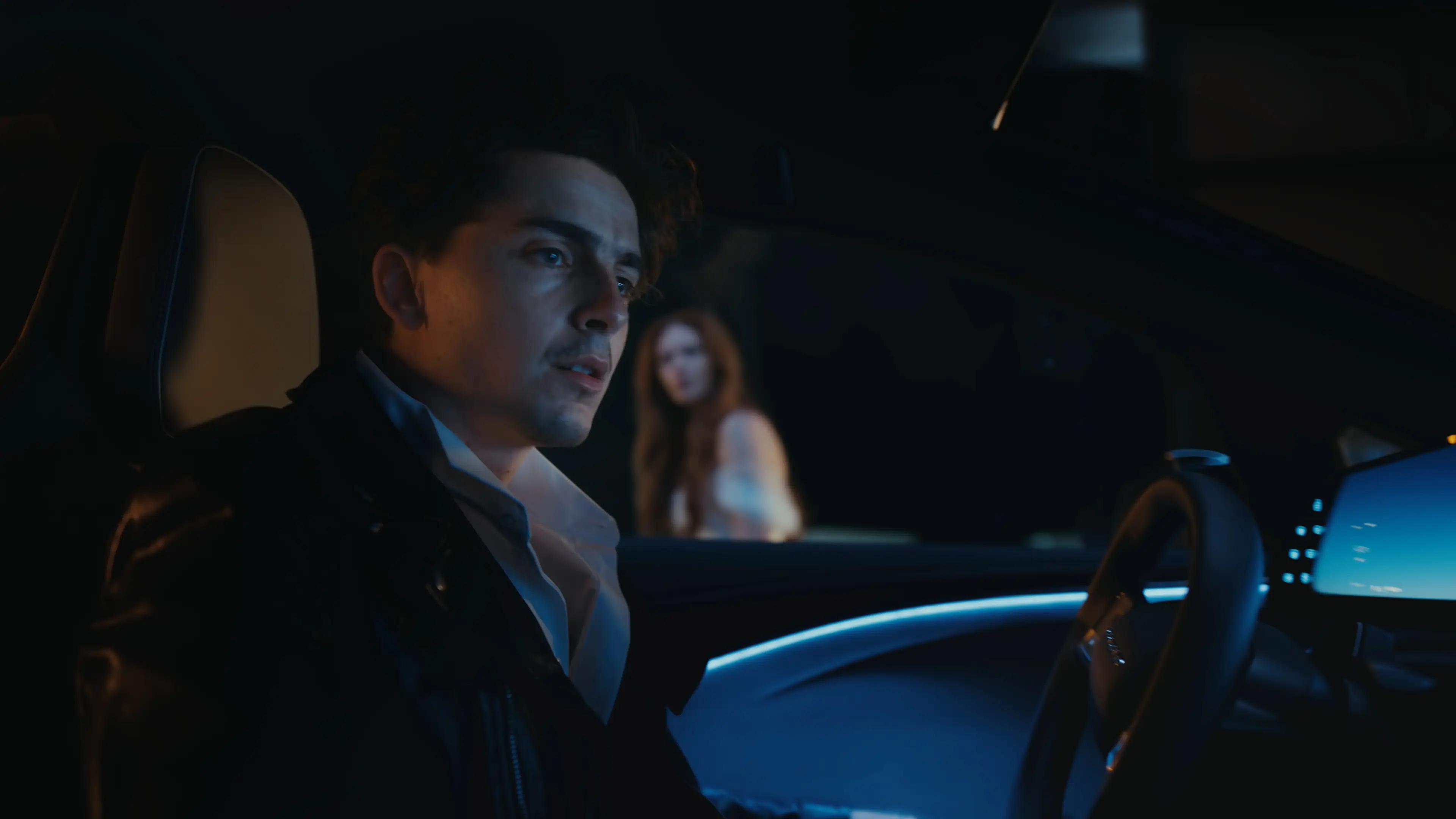 Timothée Chalamet in a jacket sitting in a dark car interior with blue lighting, another figure visible in the background.