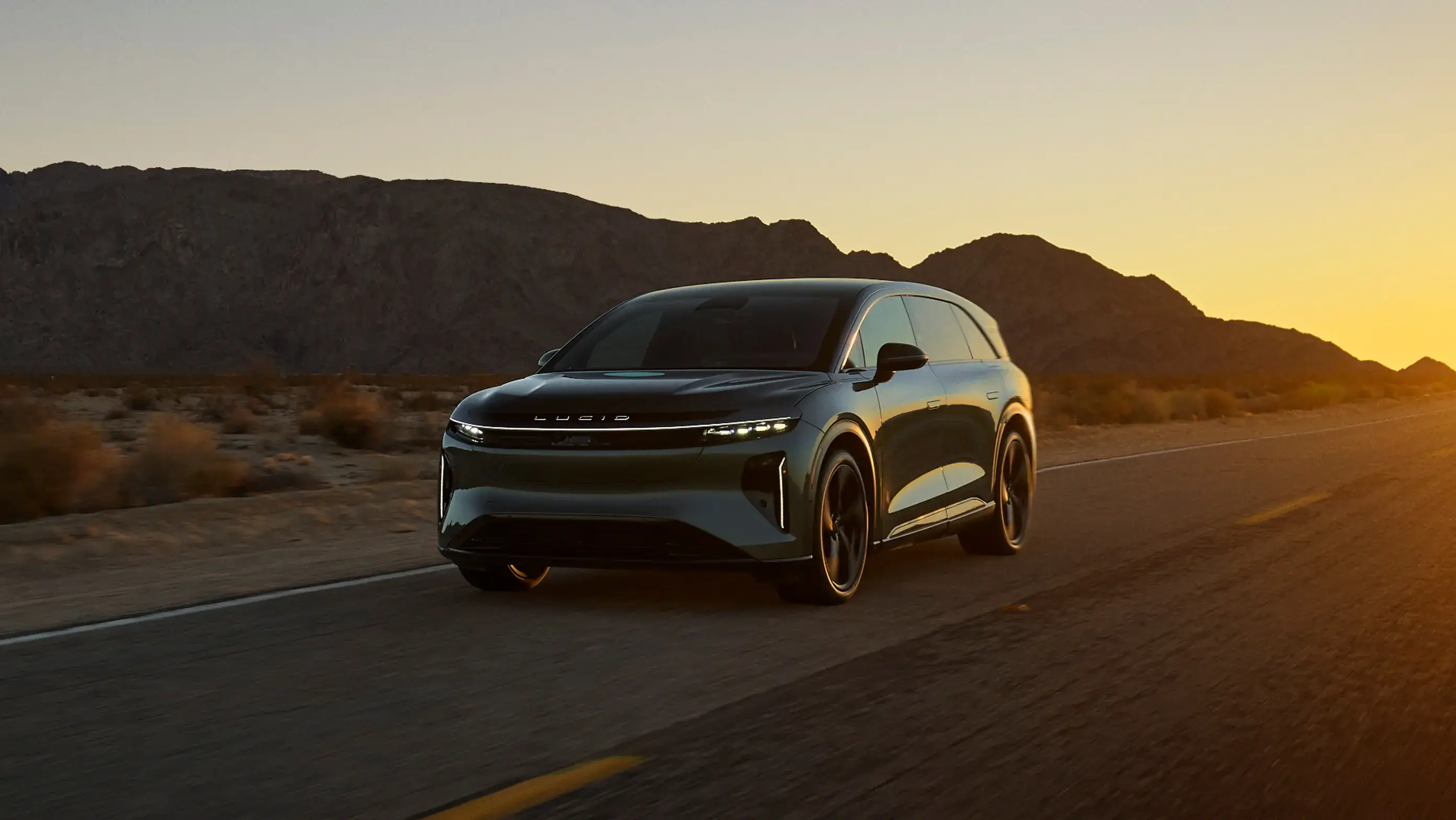 Sleek Lucid Gravity SUV driving on desert road at sunset with mountains silhouetted in the background.