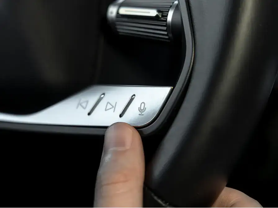 Close-up of a finger pressing media control buttons on a car's steering wheel with skip, play, and microphone icons.