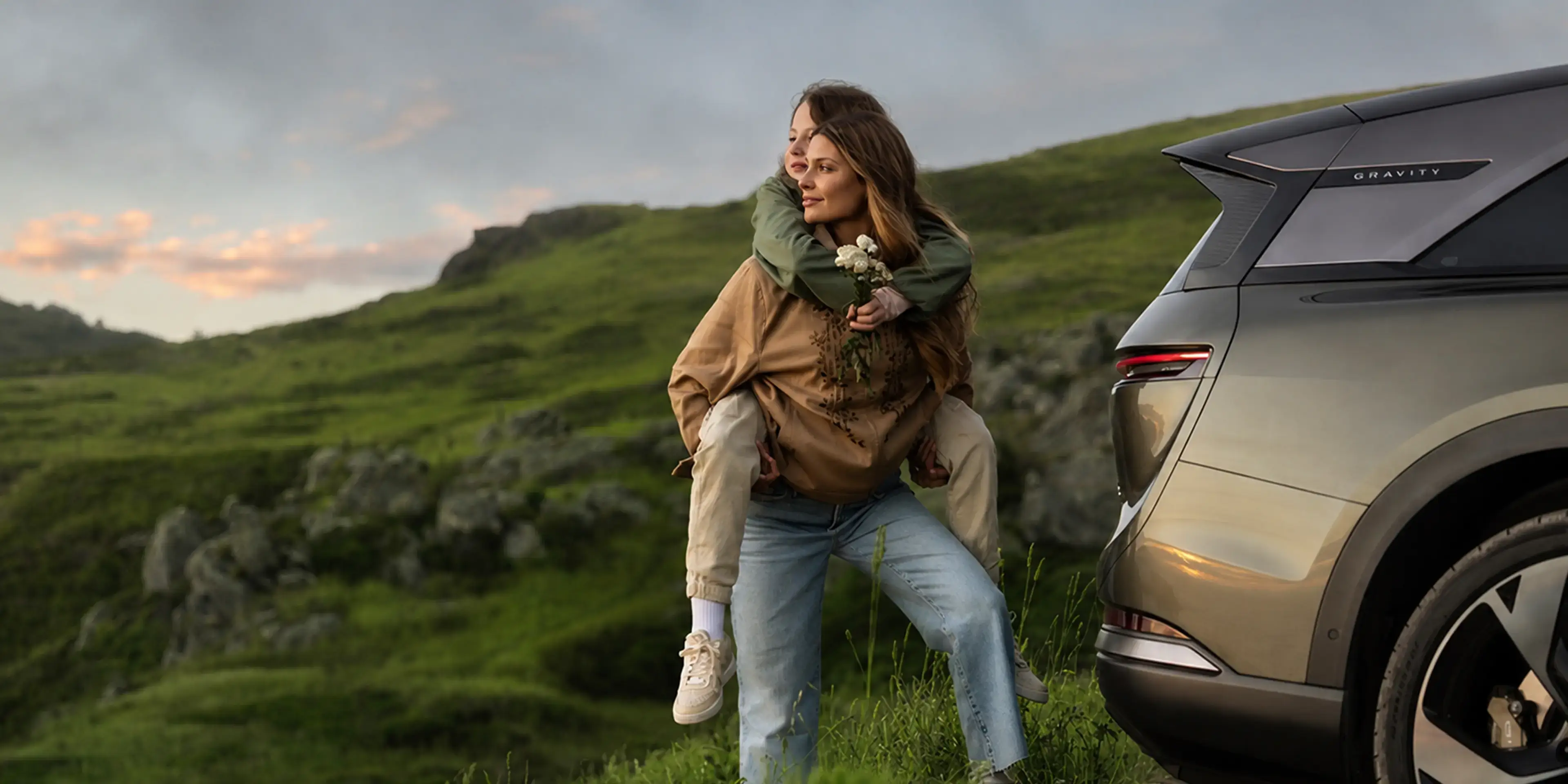 A woman gives a piggyback ride on a green hillside near a Lucid Gravity SUV at dusk, both smiling joyfully.