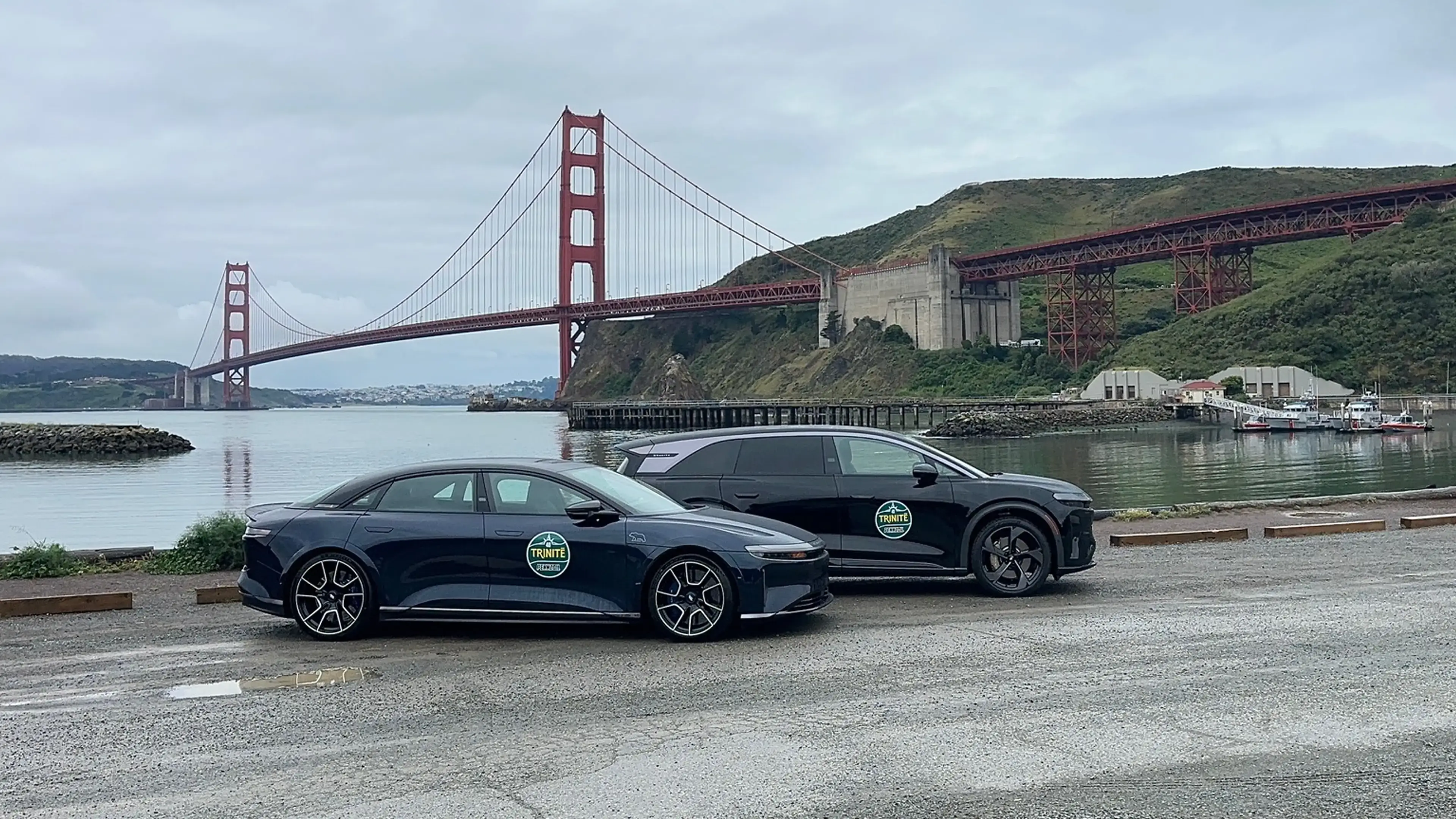 Two dark Trinité rally cars parked near the Golden Gate Bridge in San Francisco on a cloudy day.