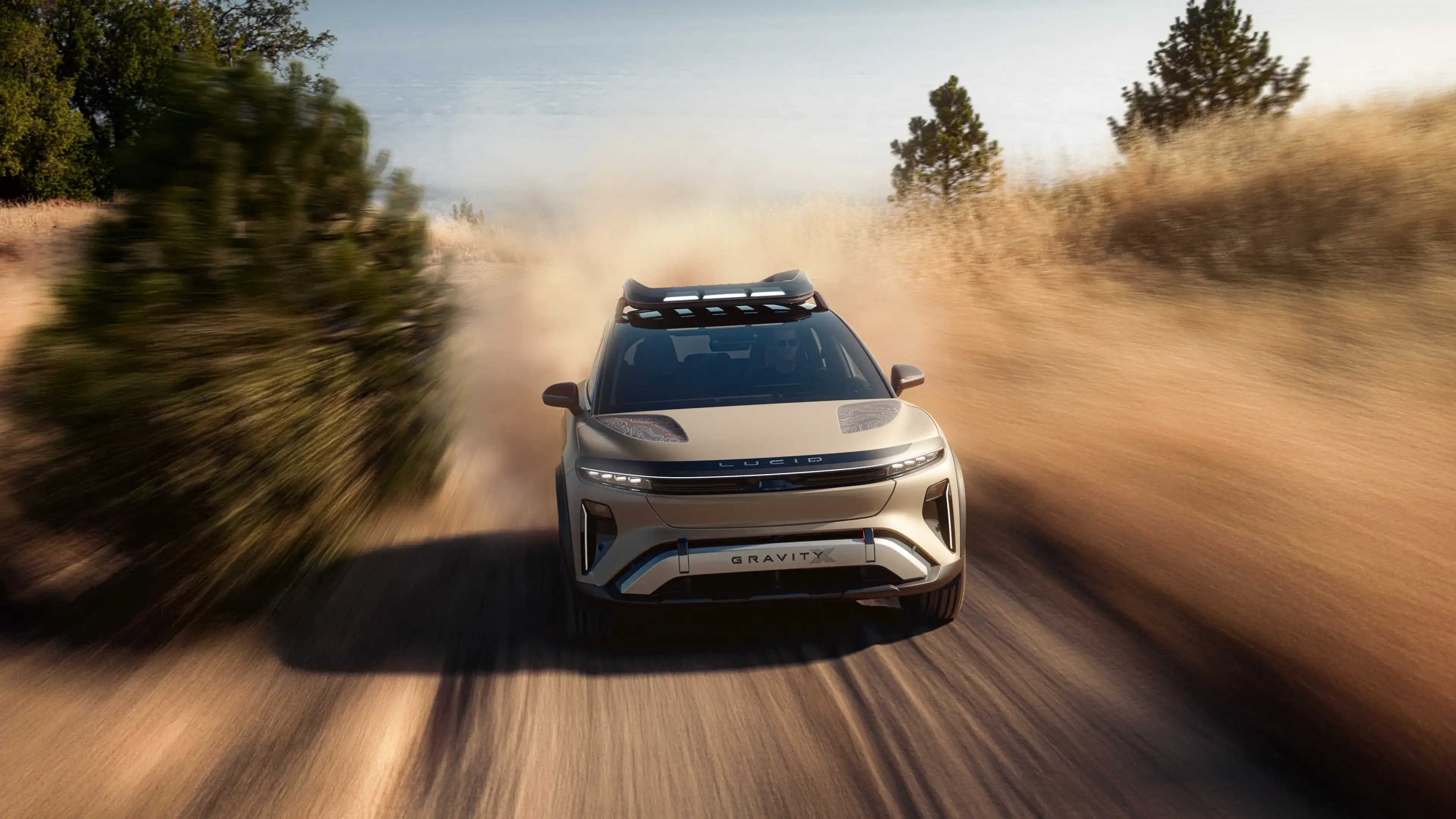 Lucid Gravity SUV speeding down a dusty dirt road, kicking up clouds of dust with pine trees on either side.