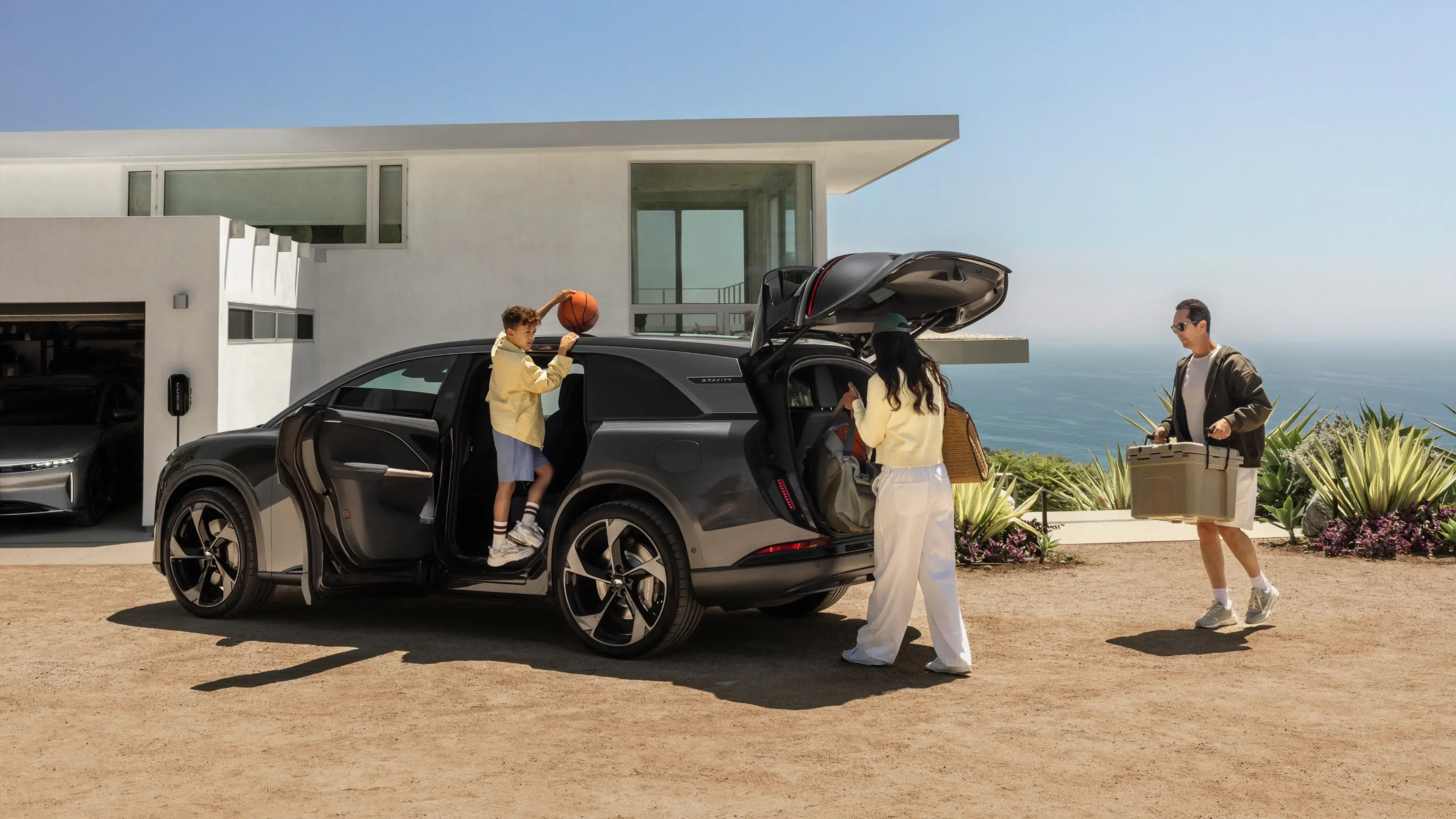 Family loading a dark gray SUV outside a modern coastal home; boy holds basketball, woman loads trunk, man carries cooler.
