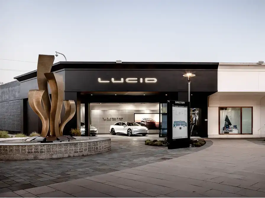 Lucid Motors showroom with illuminated logo, white electric cars on display, and a bronze sculpture outside.