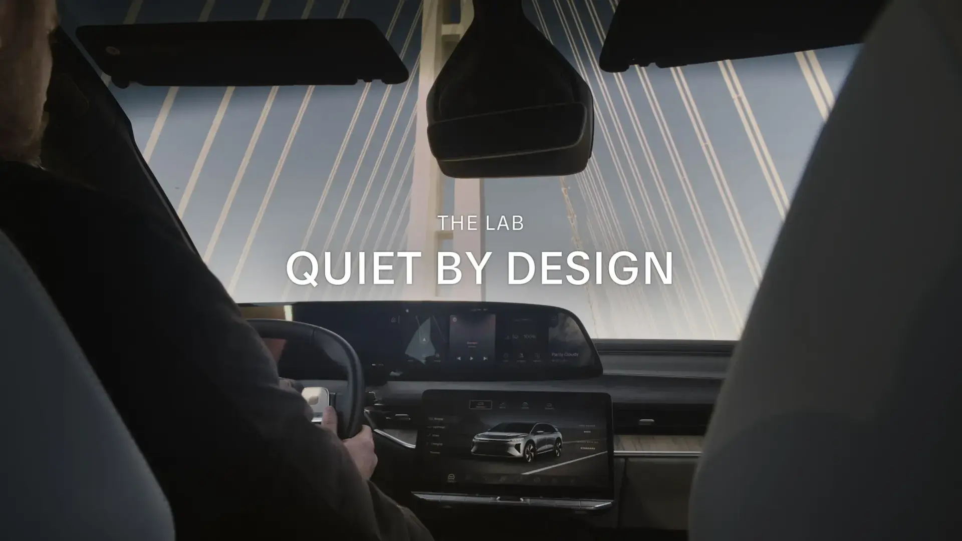 Car interior view with text "THE LAB QUIET BY DESIGN" overlaid, showing dashboard displays and bridge cables outside.