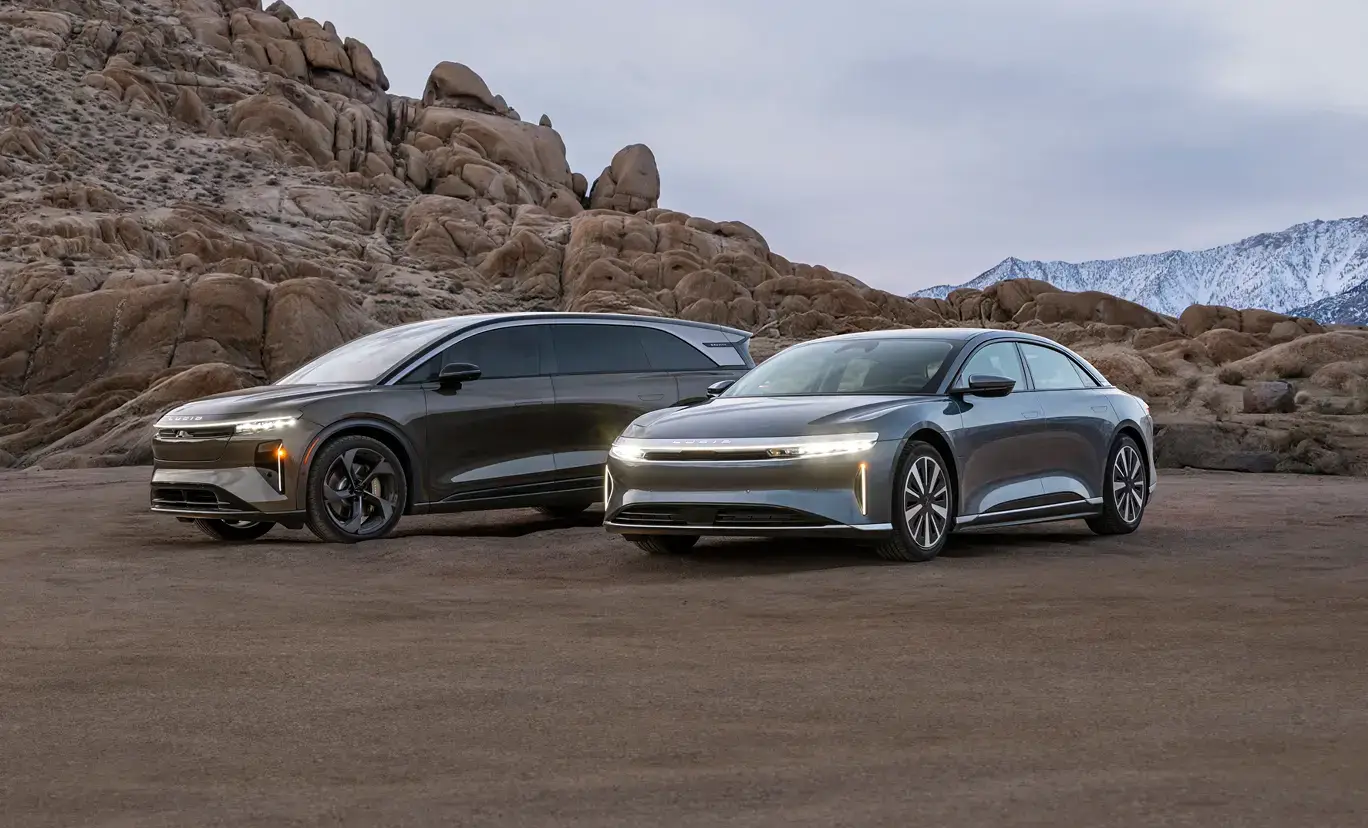 Two sleek Lucid electric vehicles parked in a desert landscape with rocky formations and snow-capped mountains in the background.