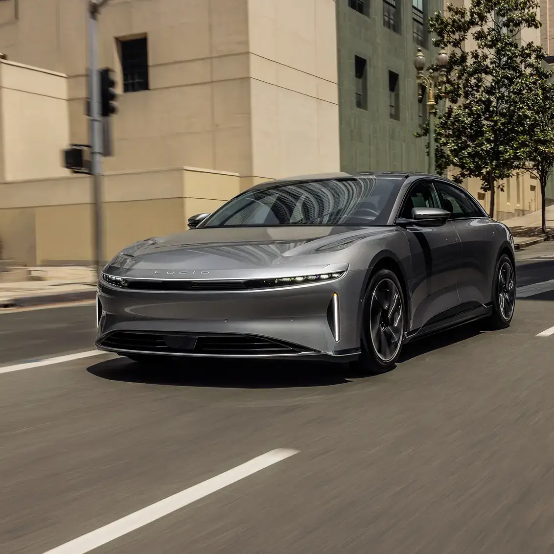 A silver Lucid Air electric sedan driving on an urban street, with buildings and a tree in the background.