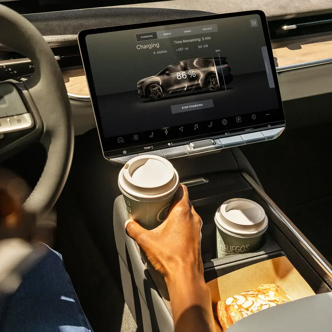 Electric vehicle dashboard showing 86% charge with 5 minutes remaining, while a hand holds coffee in the cupholder.