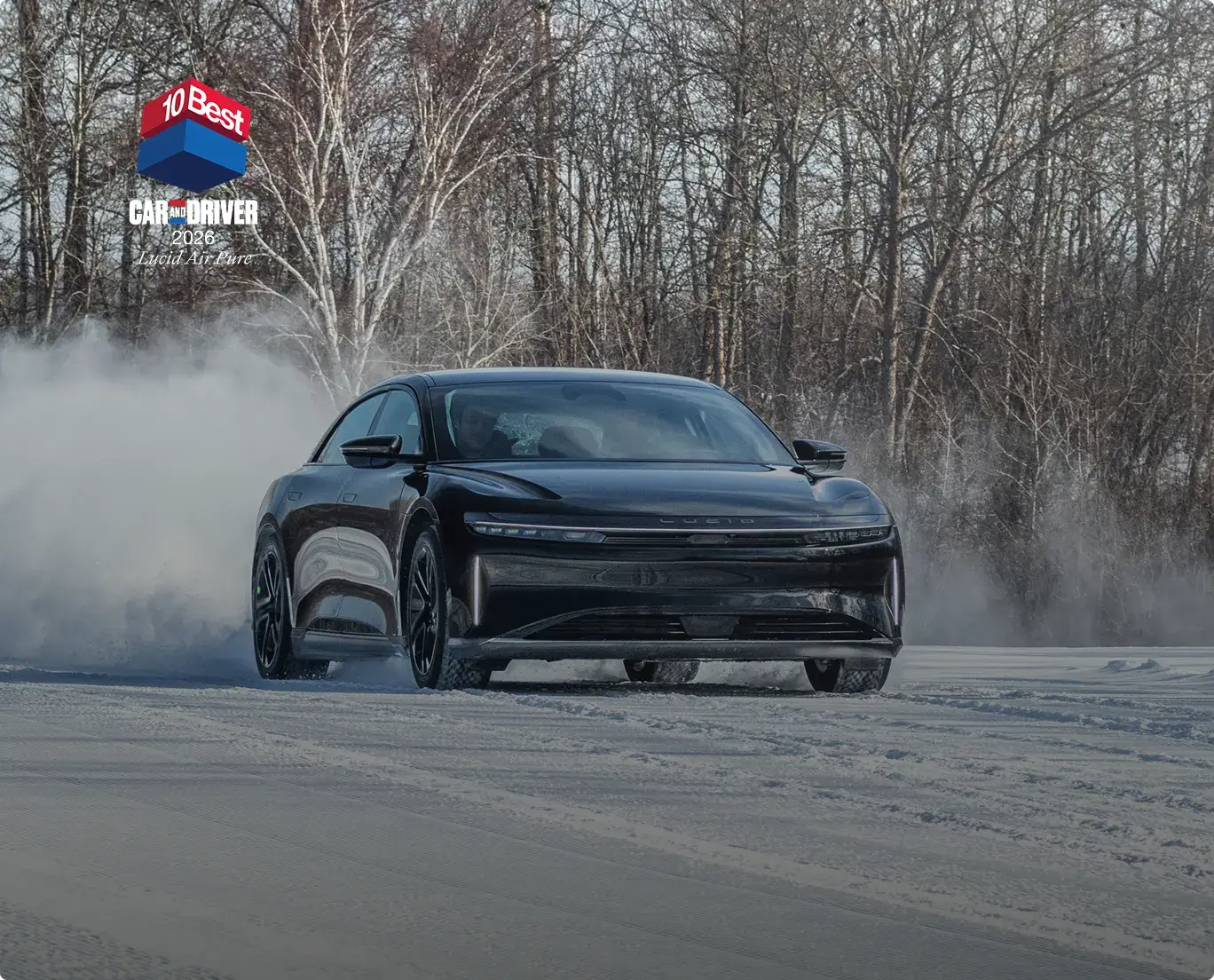 Black Lucid Air electric sedan drifting on snowy road with smoke, bare winter trees in background, Car and Driver award badge.