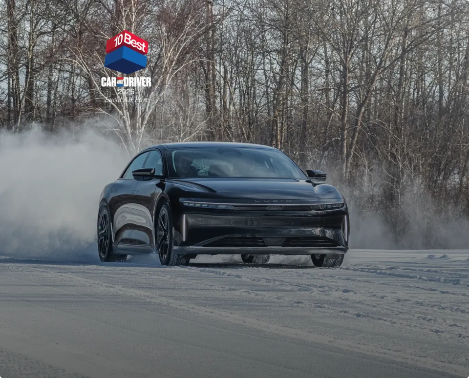 Black Lucid Air electric sedan drifting on snowy road with smoke, bare winter trees in background, Car and Driver award badge.