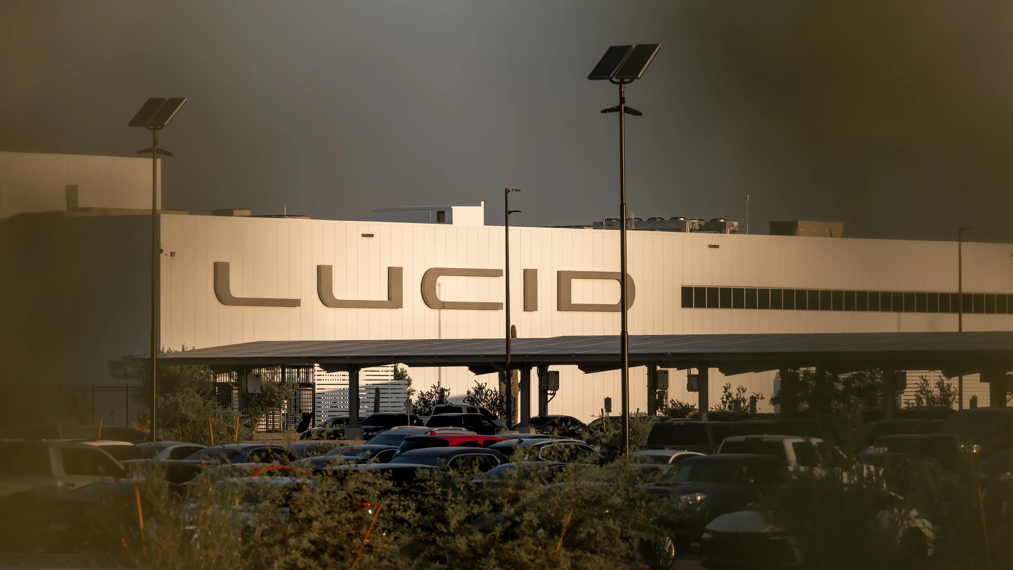 Lucid Motors factory building with large logo at sunset, parking lot with vehicles in foreground and solar-powered lights.