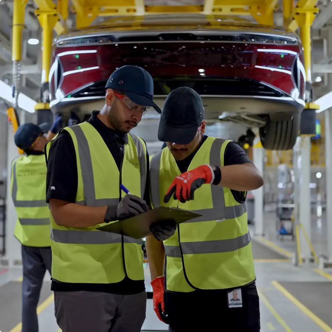 Factory workers in safety vests reviewing documents beneath a red car on an assembly line with yellow support structures.