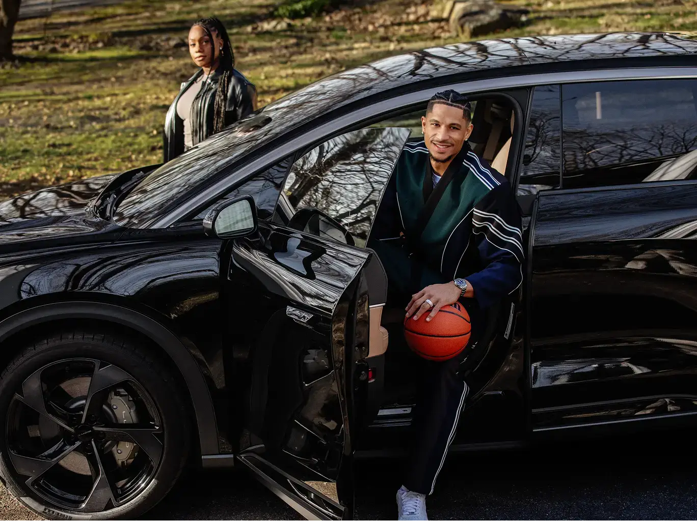 Josh Hart holding a basketball steps out of a black SUV, while a woman stands behind the vehicle outdoors.