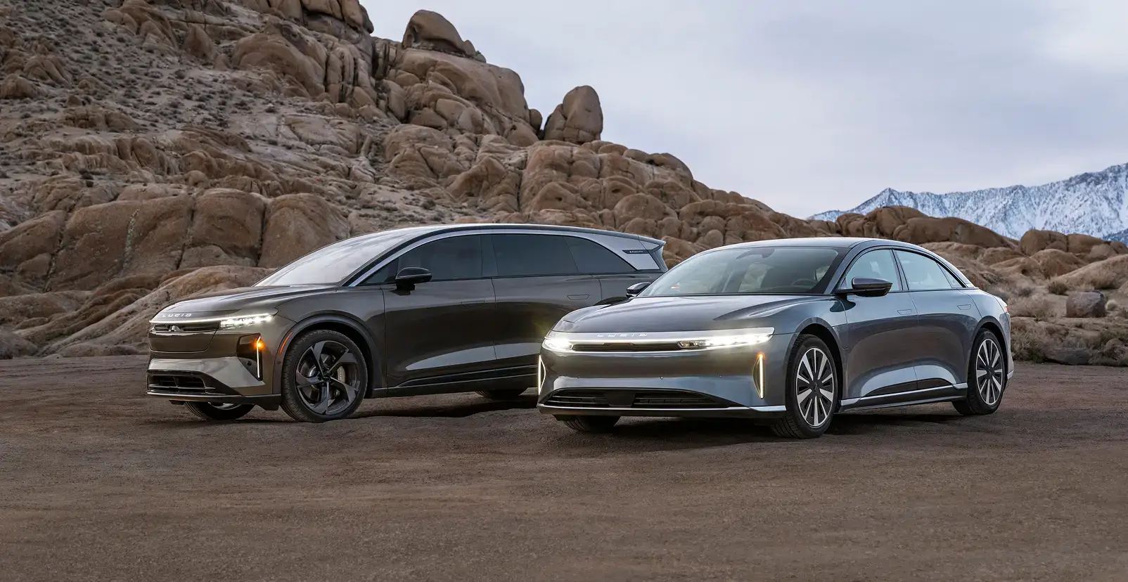 Two Lucid electric vehicles parked in a desert landscape with rocky formations and mountains in the background.