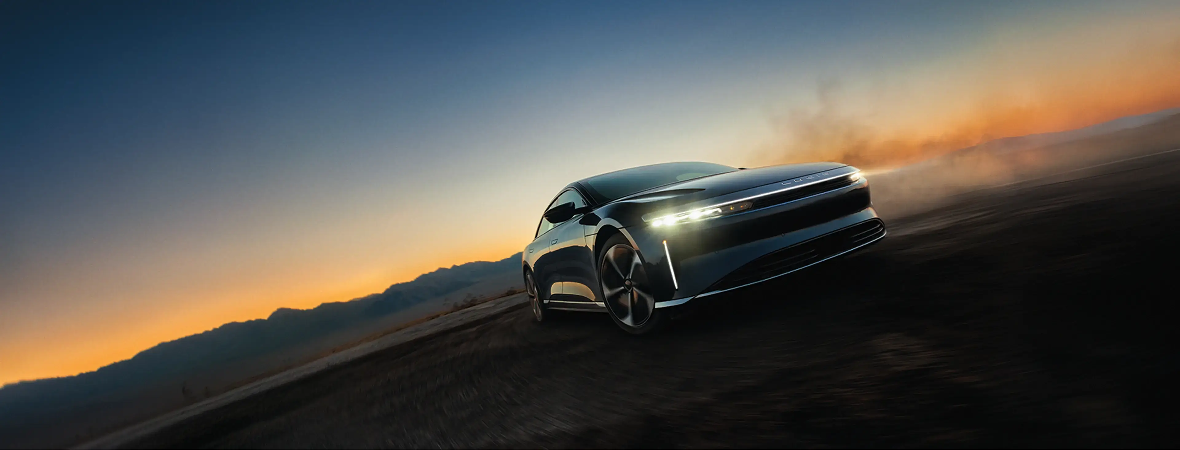 Sleek electric sedan speeding across desert terrain at sunset, headlights illuminated against an orange and blue sky.