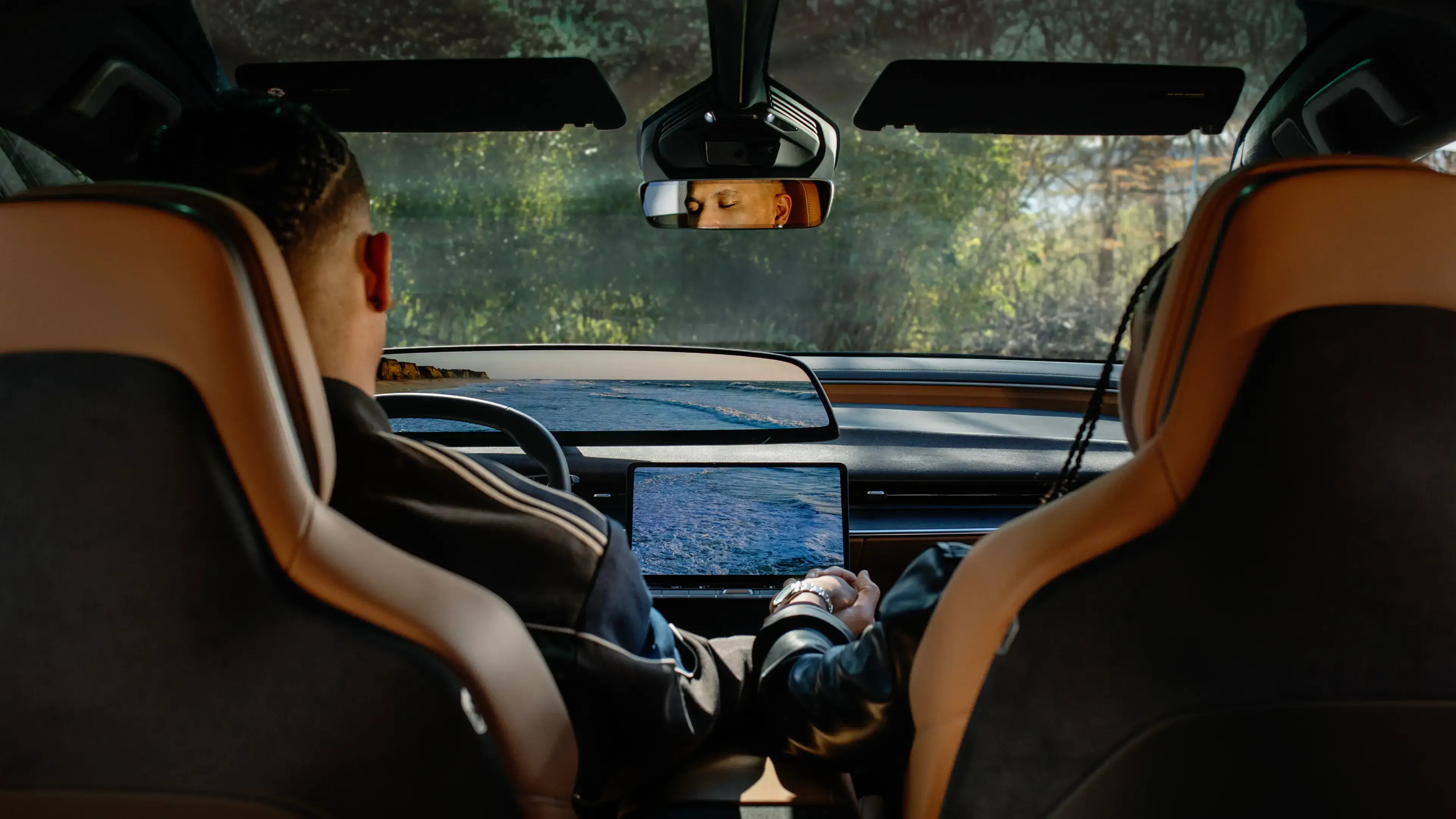Person sitting in a car's front seat using a touchscreen dashboard, viewed from the back seat with tan leather interior.