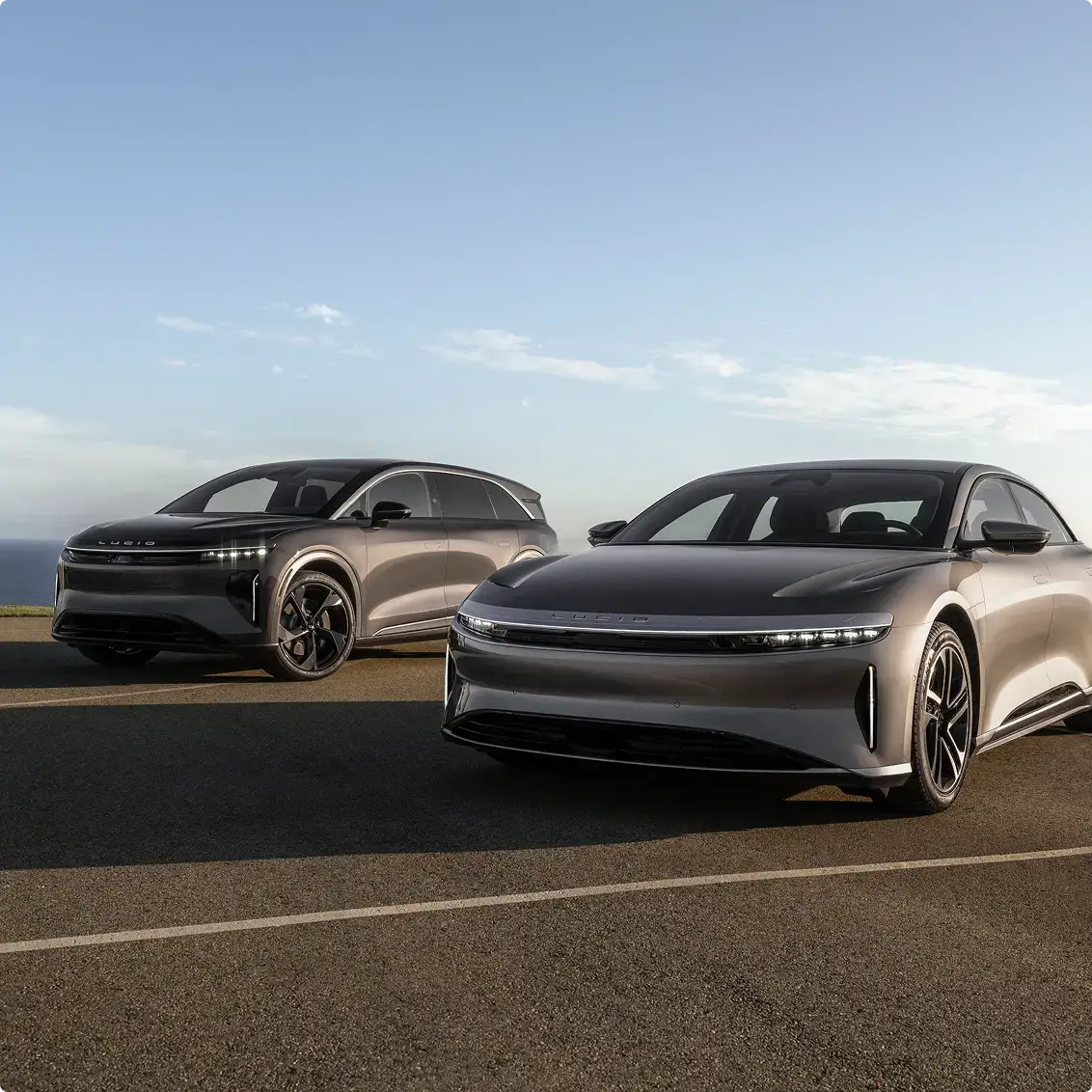 Two Lucid electric vehicles — a sedan and an SUV — parked on a road under a clear blue sky.
