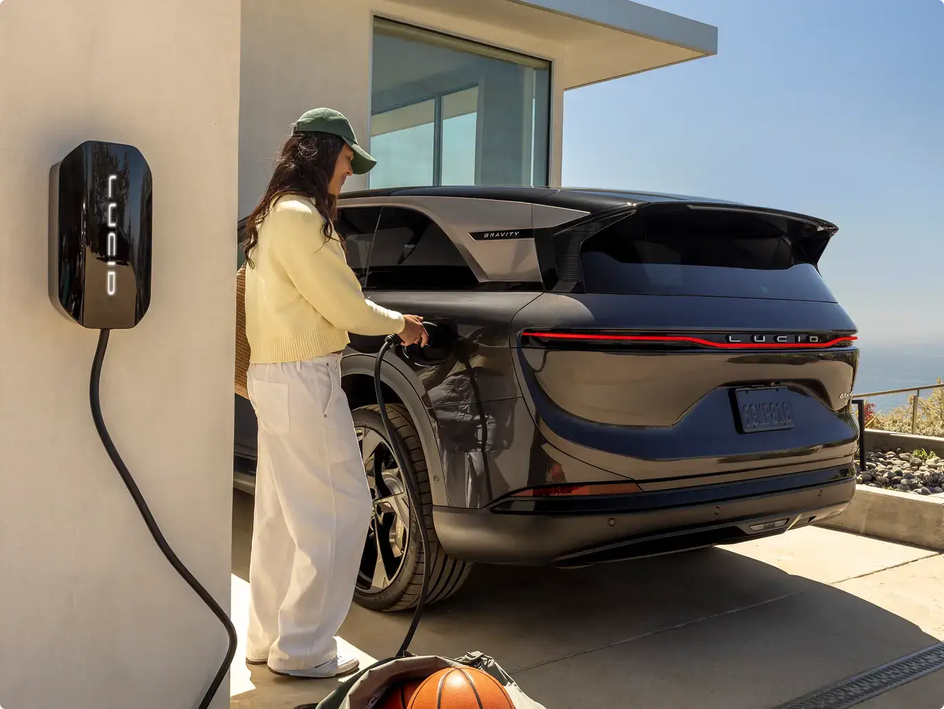 Woman in yellow jacket charging a dark blue electric SUV at a home Lucid charging station with ocean view.