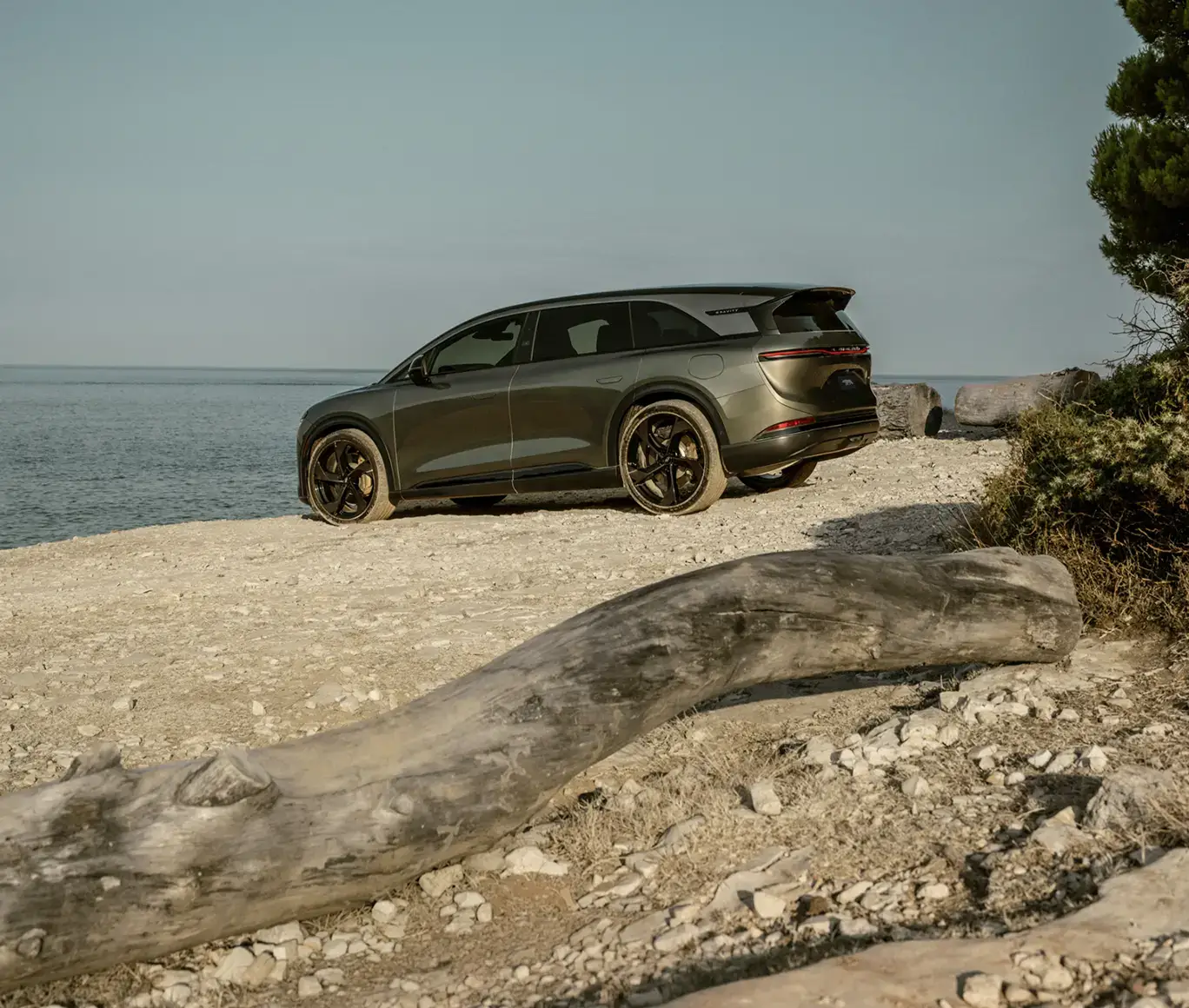 Dark olive green luxury SUV parked on a rocky shoreline with driftwood, calm water, and a clear sky in the background.