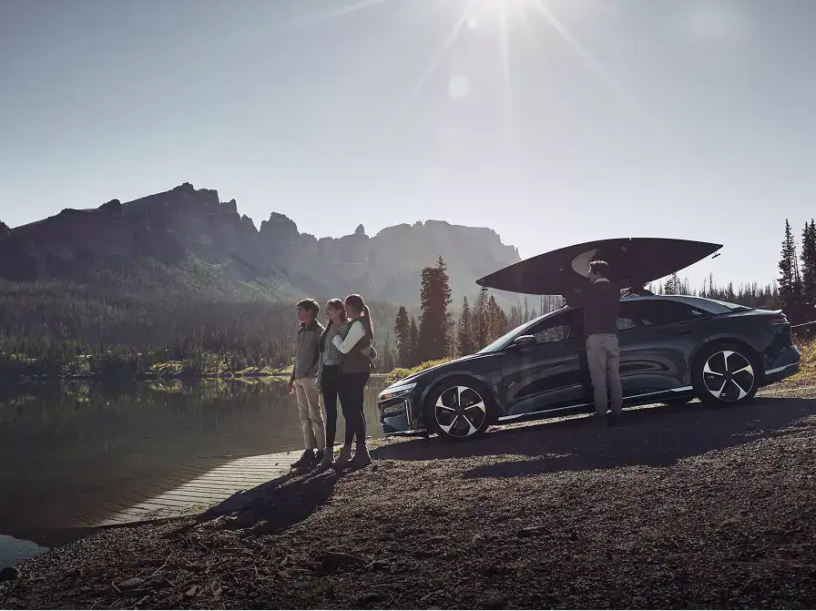 Family of three standing by a lake near a Luid Air with a kayak mouunted on top, mountains and trees in background.