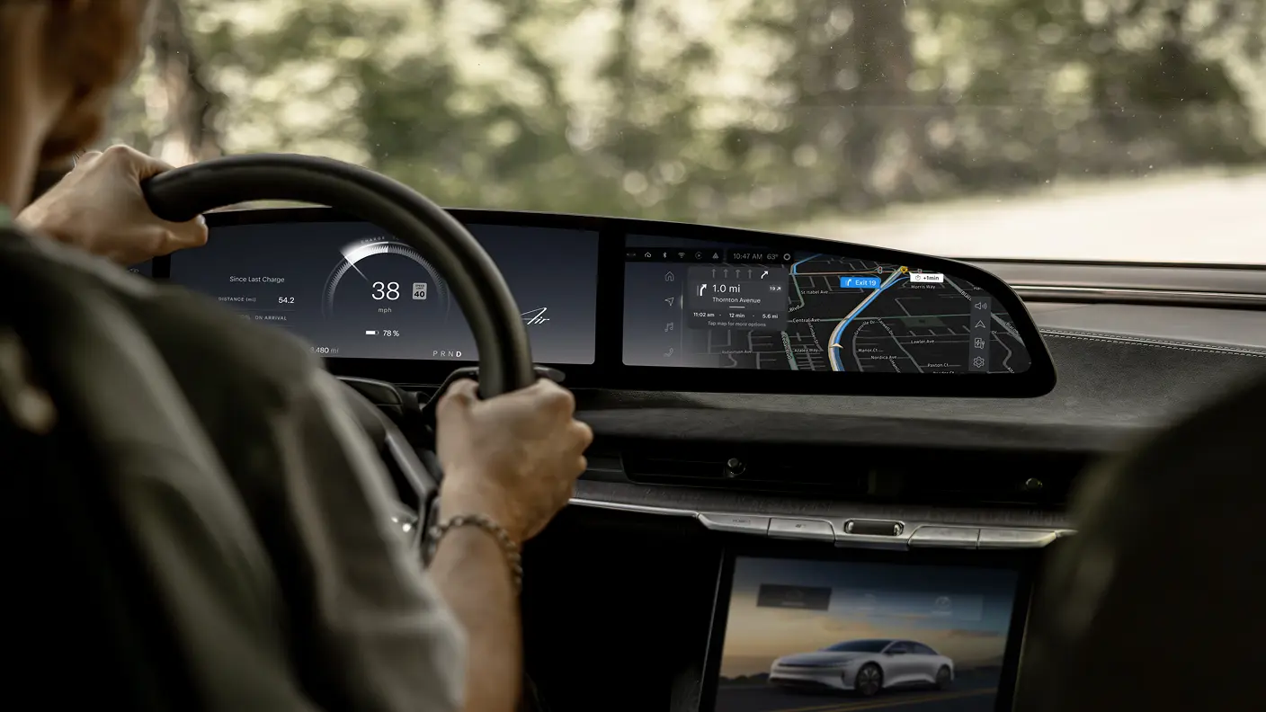Driver gripping steering wheel with wide dashboard display showing speedometer, navigation map, and a white electric car on a center screen.