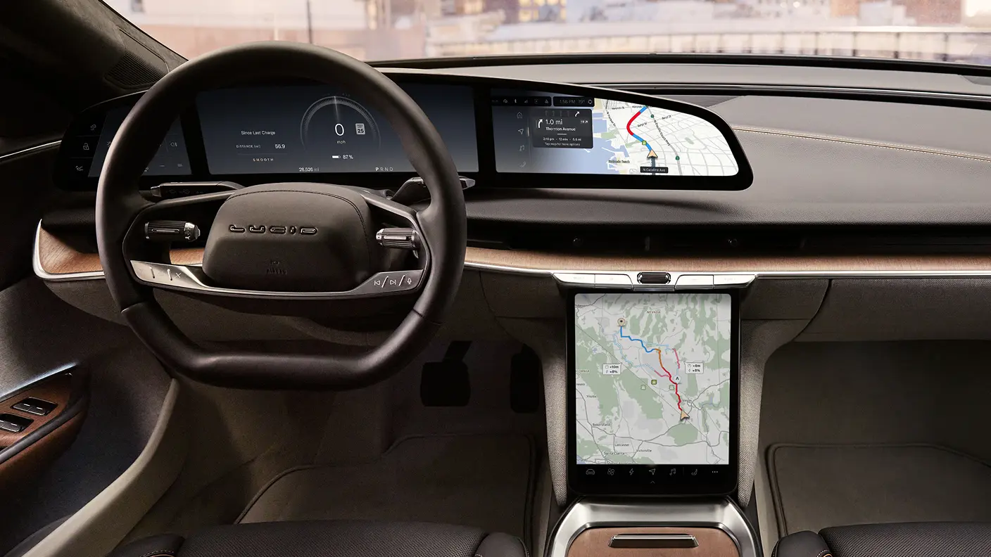 Driver gripping steering wheel with wide dashboard display showing speedometer, navigation map, and a white electric car on a center screen.