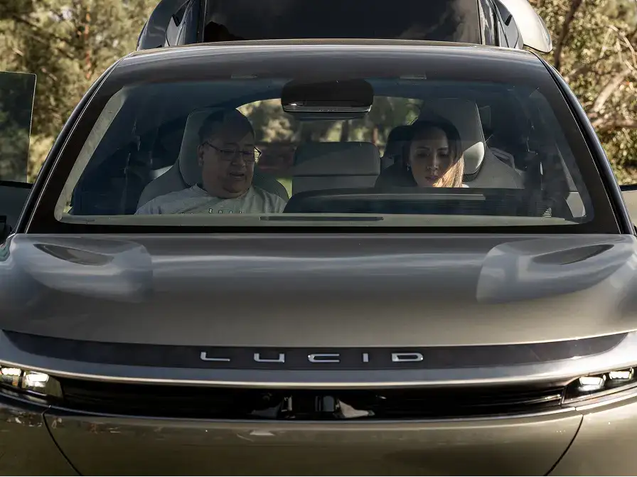 Two people sitting inside a gray Lucid electric vehicle, viewed from the front, with trees in the background.
