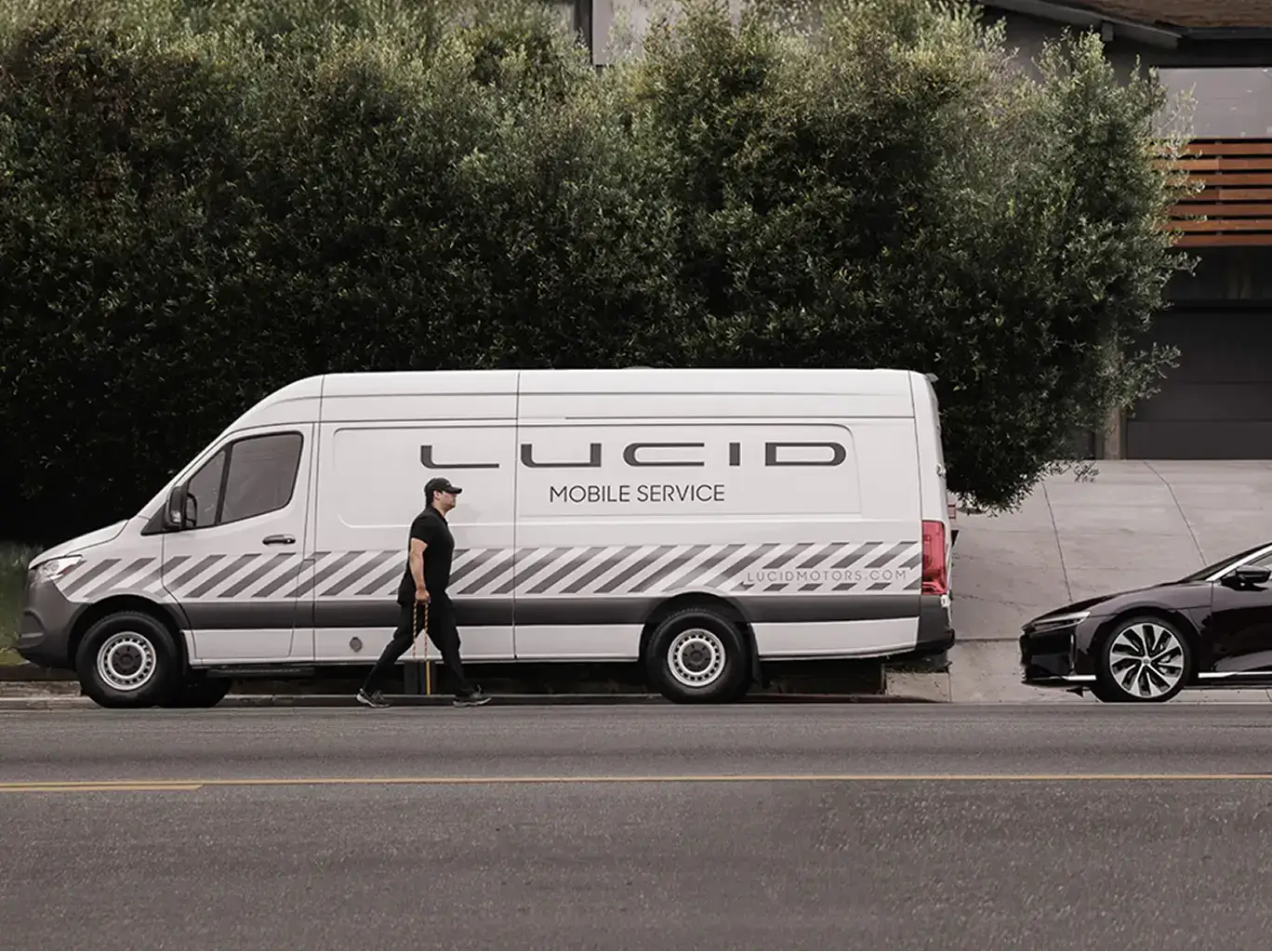 White Lucid Mobile Service van parked on street with technician walking alongside, green hedges in background.