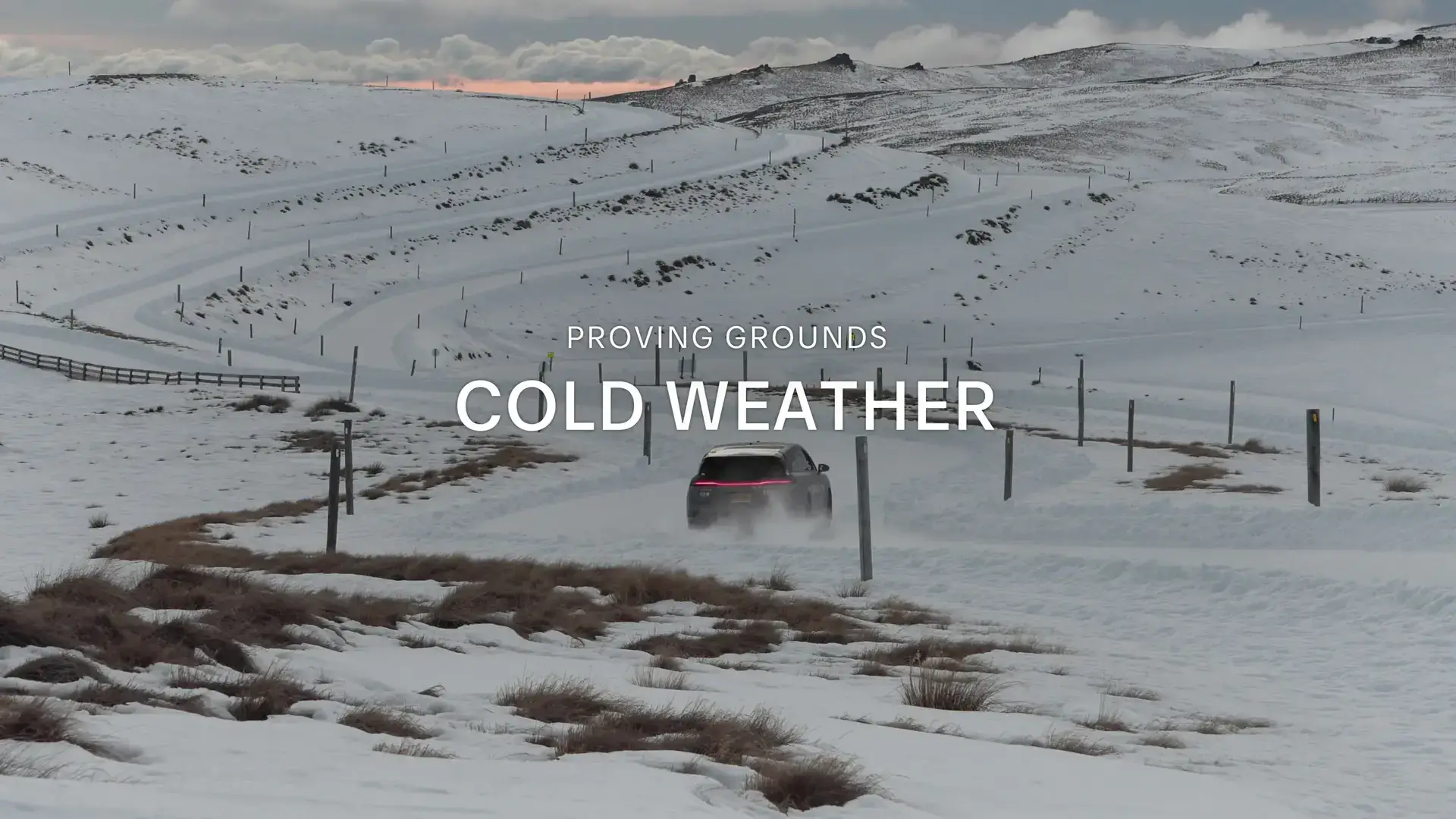 Car driving through snowy proving grounds with text overlay "PROVING GROUNDS COLD WEATHER" against winter landscape.