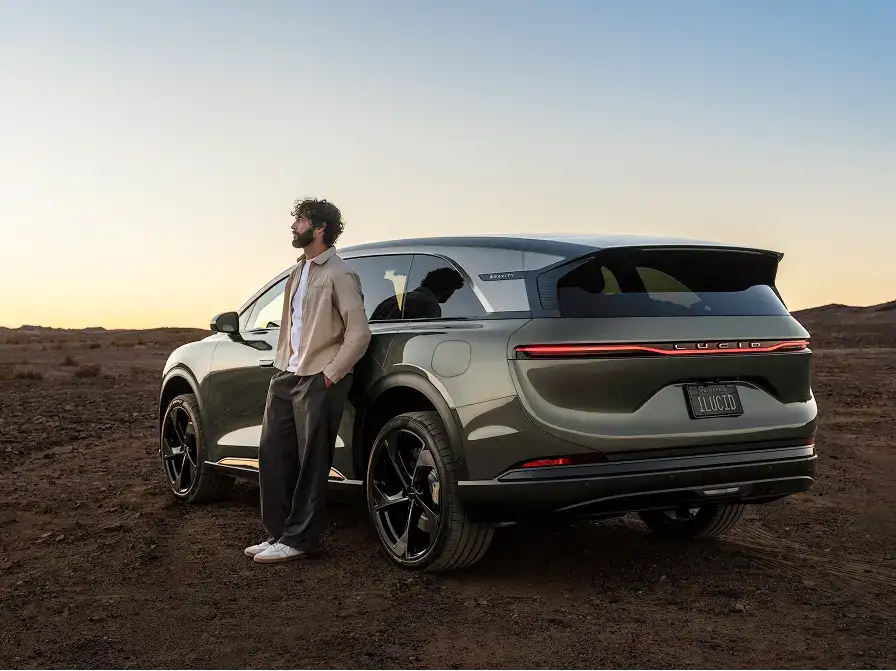 A man leans against a dark green Lucid Gravity SUV in a desert landscape at sunset, gazing into the distance.